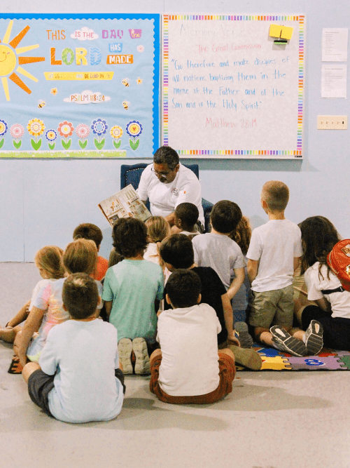 Arkansas Chef Reading To Young Kids A teacher sits on a chair reading a book to a group of young children seated on the floor, facing colorful classroom bulletin boards with religious messages and artwork.