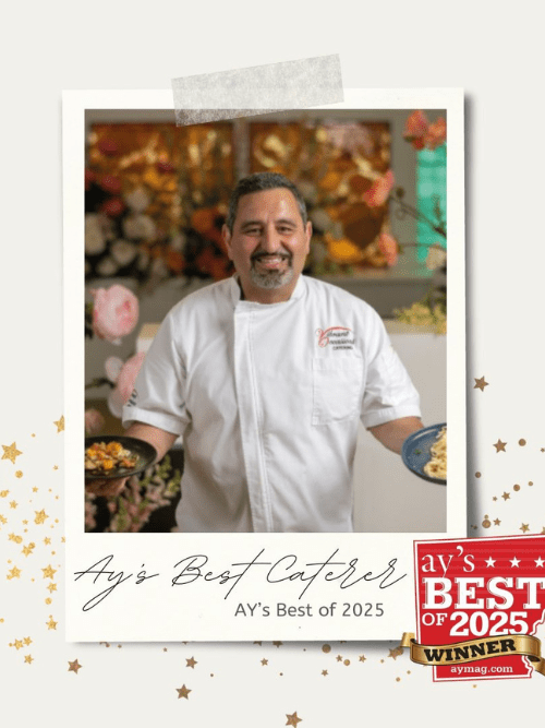 Award Winning Caterer A smiling chef in a white coat holds two plates of food. The photo has a decorative border with gold stars and text that reads “Ay’s Best Caterer, Ay’s Best of 2025 Winner.” Flowers appear in the background.