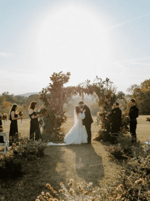 Boho Arkansas Wedding A bride and groom kiss under a floral arch at an outdoor wedding ceremony, surrounded by bridesmaids and groomsmen dressed in black, with sunlight shining brightly behind them.