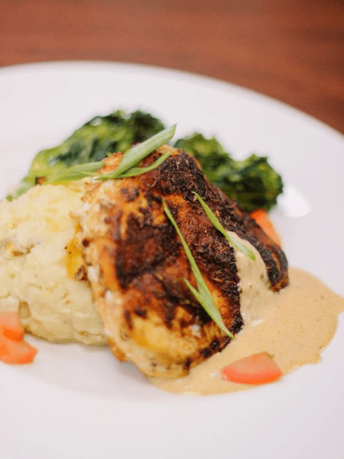 Fundraiser Plated Meal Arkansas A plate featuring a grilled, seasoned chicken breast with creamy sauce, served alongside mashed potatoes, sautéed greens, and small pieces of carrots, garnished with sliced green onions.