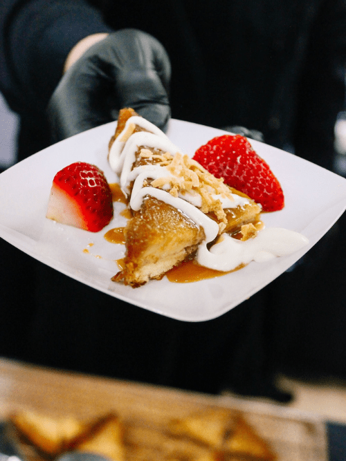 OMG French Toast Vibrant Occasions Catering A person wearing a black glove holds a plate with caramel-drizzled bread pudding, two strawberry halves, whipped cream, and toasted coconut flakes.