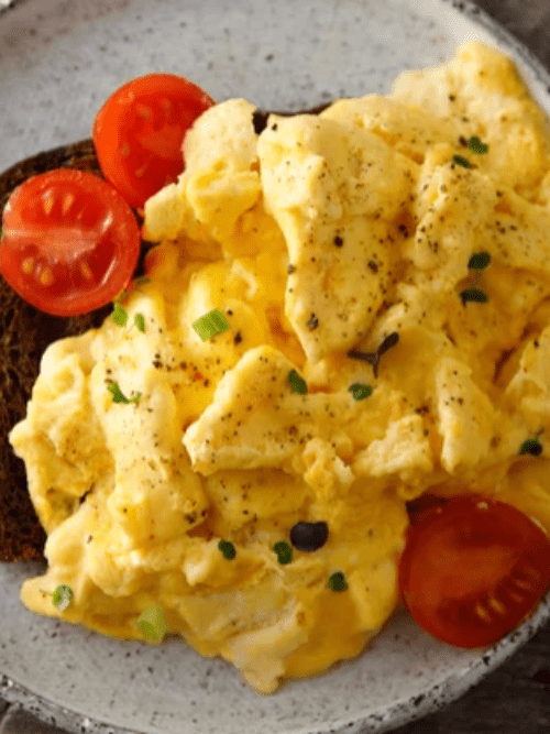 Orange Juice in Scrambled Eggs A plate of scrambled eggs garnished with chopped chives and black pepper, served with cherry tomato slices and a piece of brown toast.