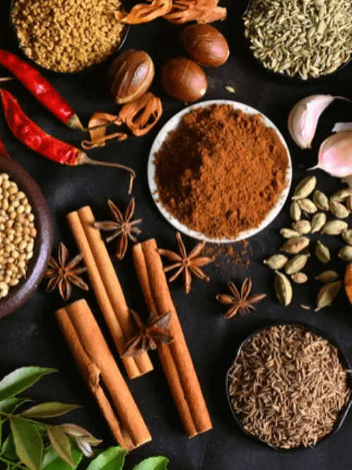 Spices Used In Arkansas Cooking An assortment of spices on a dark surface, including cinnamon sticks, star anise, nutmeg, chili peppers, coriander seeds, cumin seeds, cardamom, cloves, fennel seeds, curry leaves, garlic cloves, and ground spice powder.