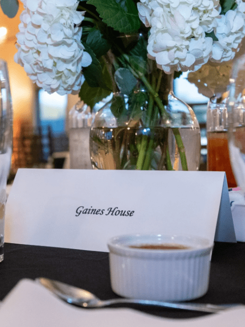 The Gaines House Arkansas A table place card reading Gaines House sits in front of a vase with white flowers, surrounded by glasses and a small white ramekin on a black tablecloth.