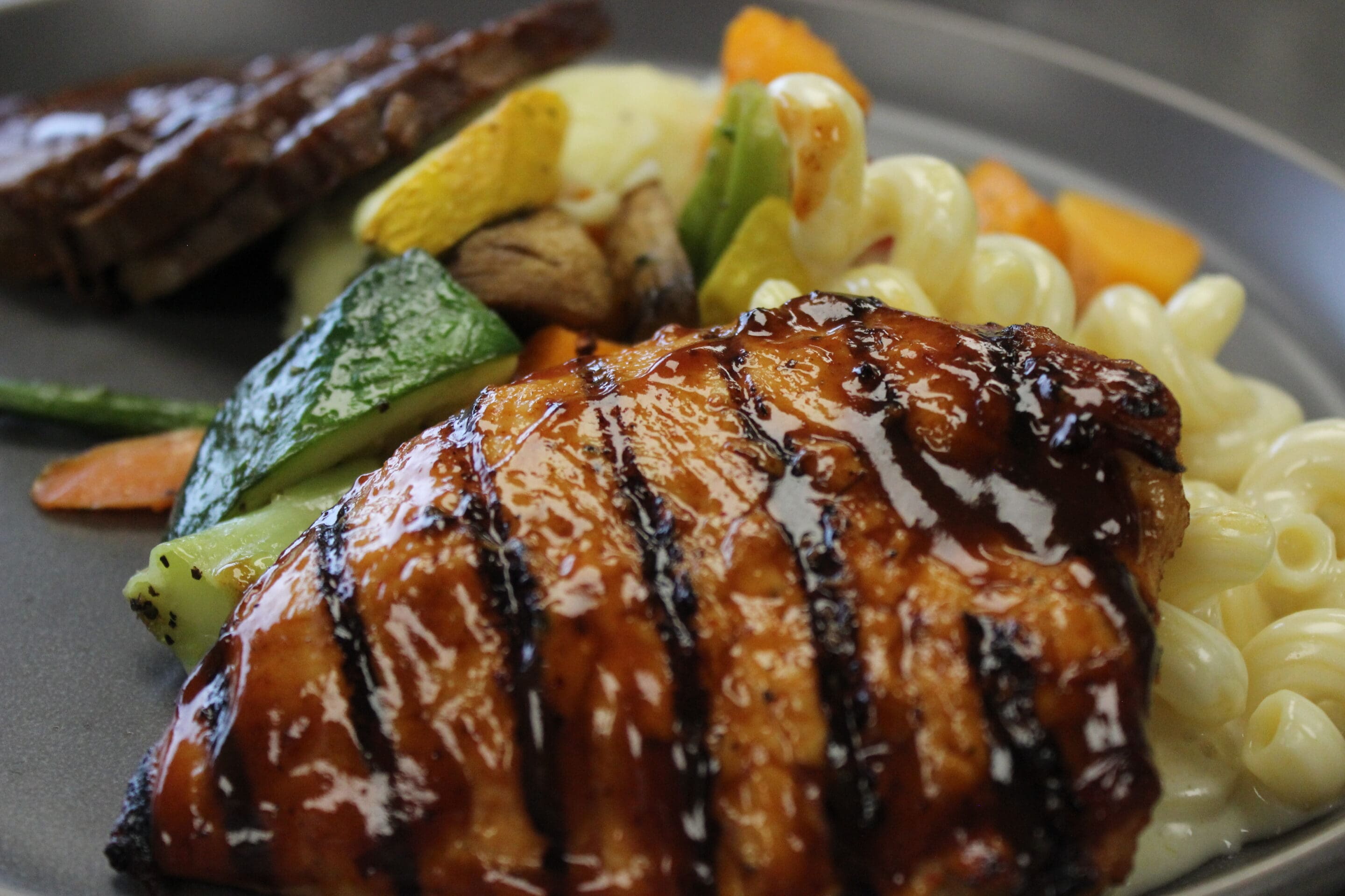 A close-up of a plate with glazed grilled chicken breast, creamy macaroni, sautéed vegetables, and a portion of steak in the background.