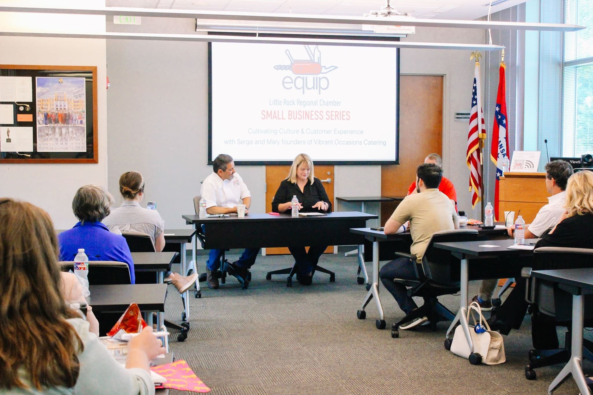 People sit at tables in a conference room, facing two speakers at the front beneath a screen displaying “Small Business Series” for the Little Rock Regional Chamber’s Equip program. American and Arkansas flags are visible.