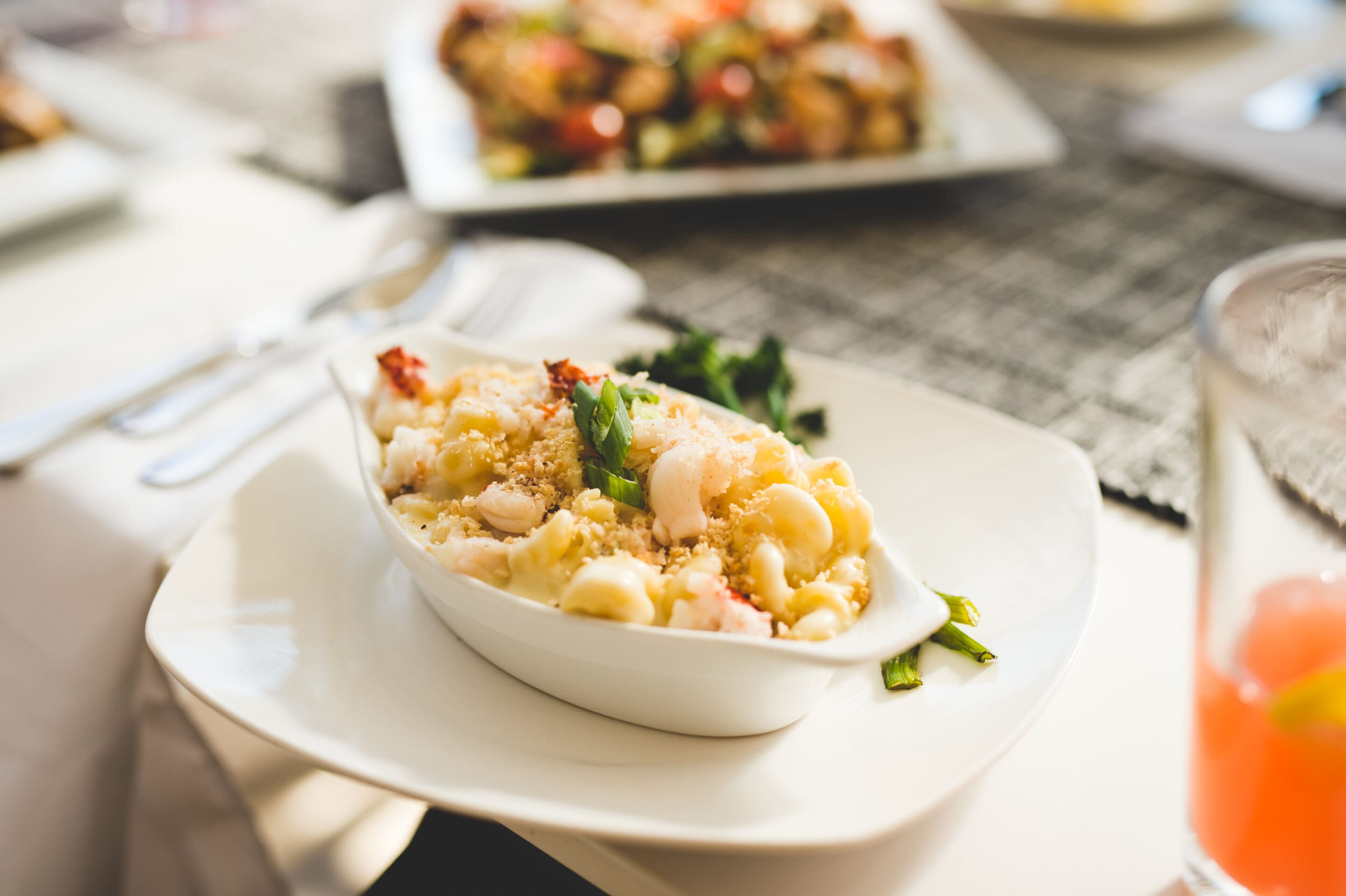 A white oval bowl filled with baked macaroni and cheese topped with breadcrumbs and herbs, served on a white plate. Blurred background shows more food and a glass with a pink drink.