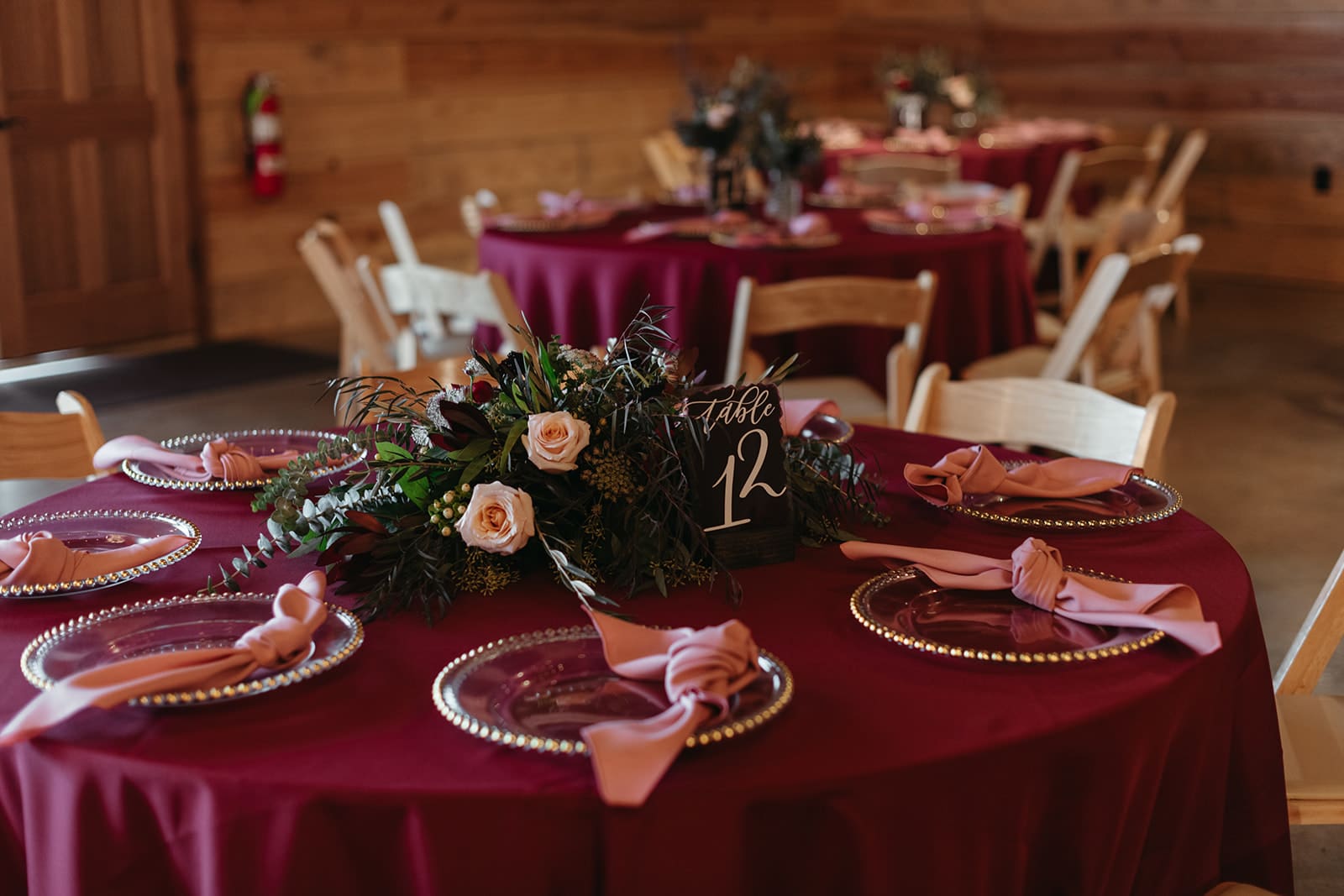 A round table with a burgundy tablecloth, set with clear plates, pink napkins, and a floral centerpiece featuring roses and greenery. A sign reads Table 12. Other similarly set tables are in the background.