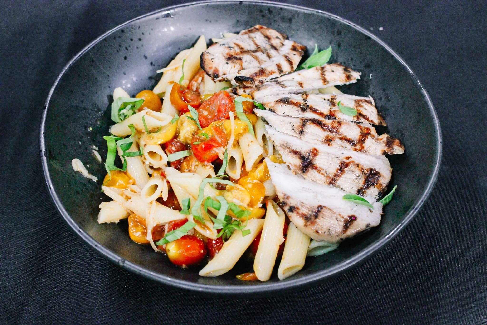A black bowl containing penne pasta with colorful cherry tomatoes and fresh herbs, served alongside sliced grilled chicken breast with visible grill marks, set on a dark background.