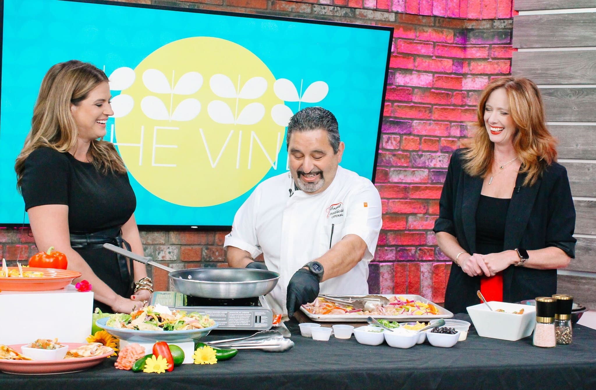 A chef smiles while cooking at a kitchen counter, surrounded by fresh ingredients, as two women stand on either side of him, laughing. A colorful sign reading THE VINE is displayed in the background.