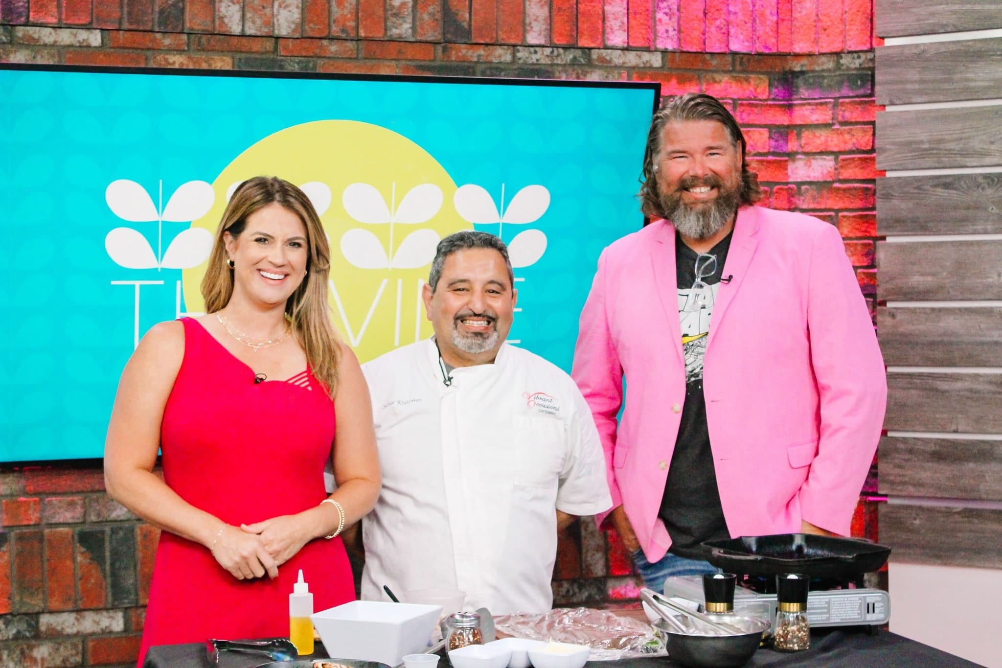 Three people smile while standing in a colorful TV studio kitchen set. A woman in a bright pink dress stands next to a chef in white, with a man in a pink blazer on the right. Cooking supplies are on the counter in front of them.