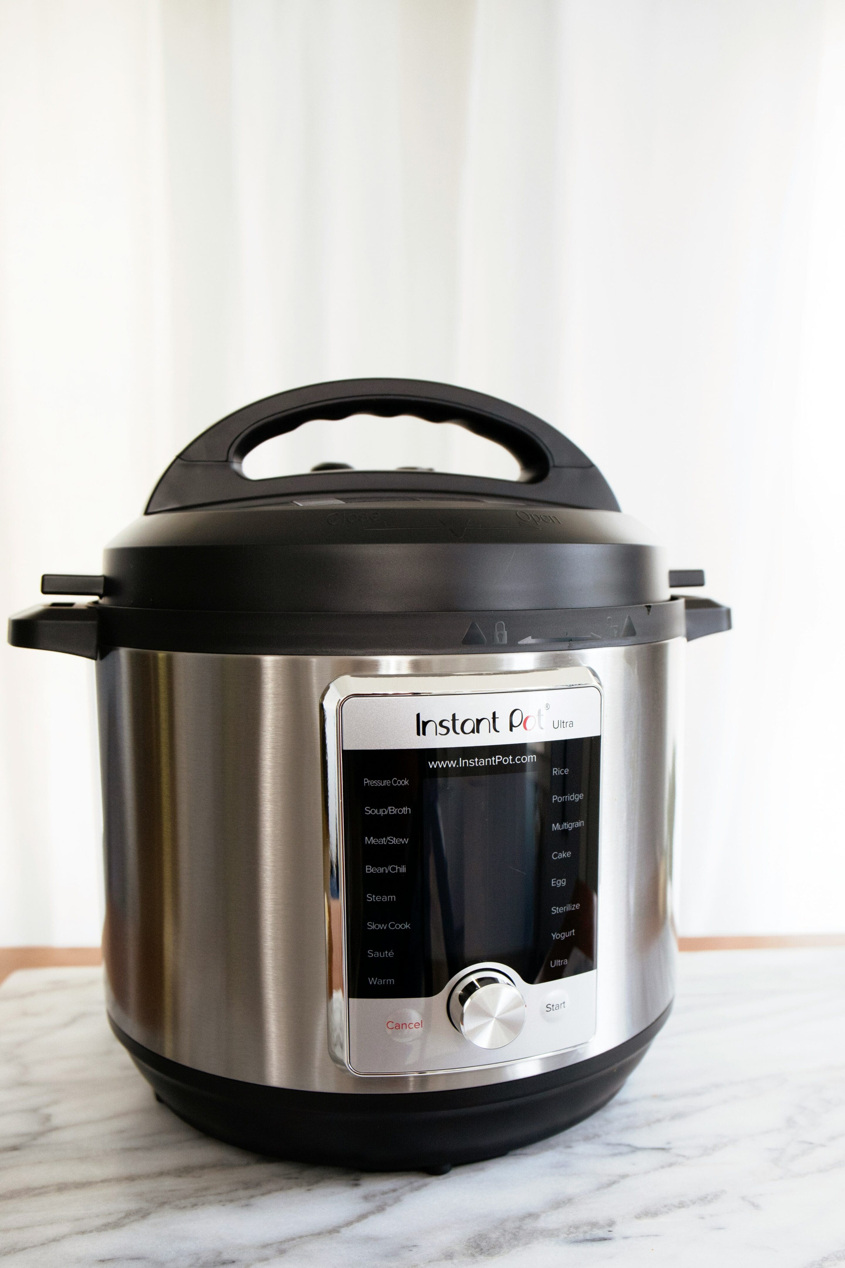 A stainless steel Instant Pot electric pressure cooker sits on a marble countertop with a white curtain backdrop. The digital control panel and dial are visible on the front.