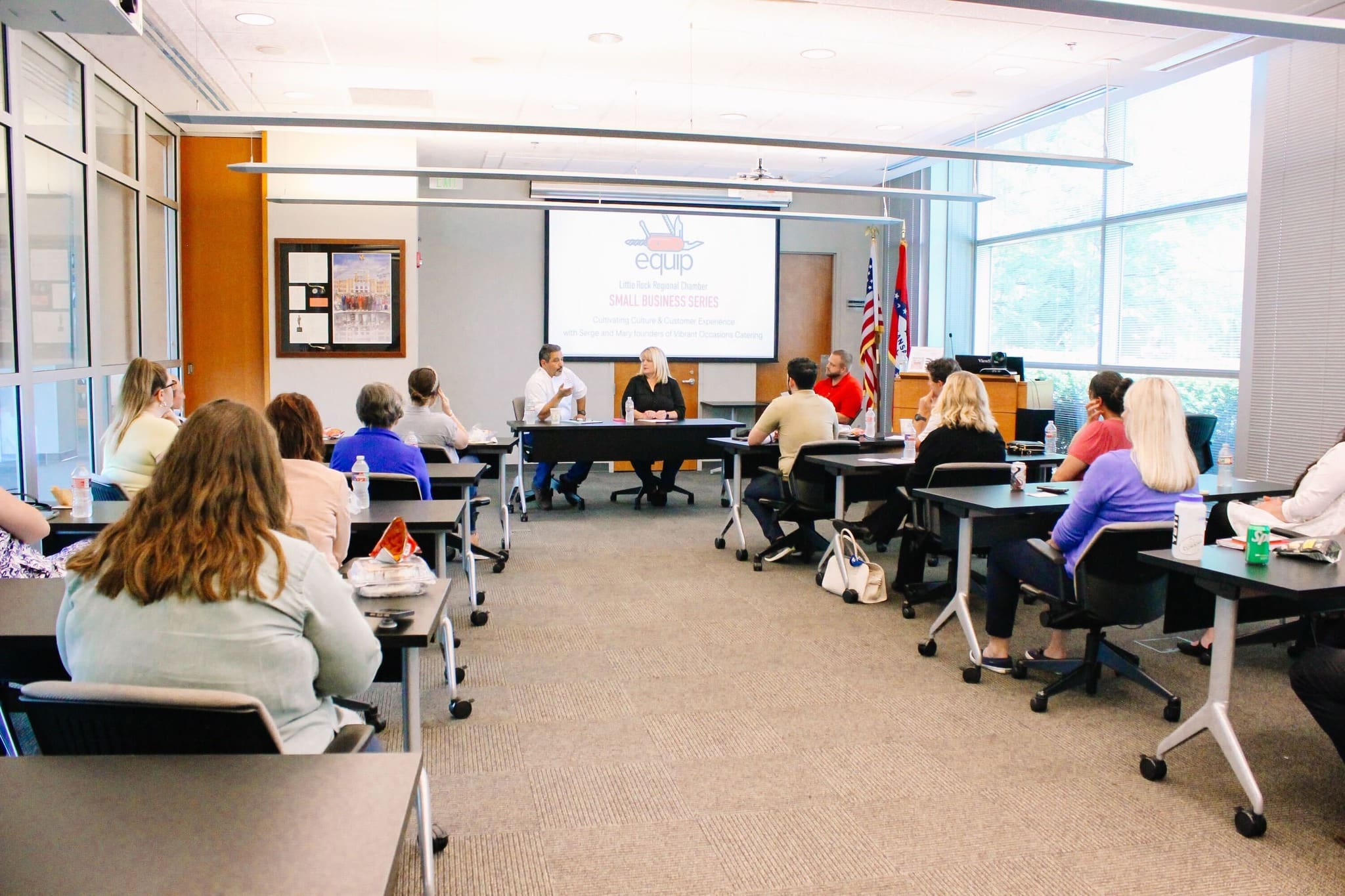 A group of people sit at tables in a conference room, facing a panel of three speakers in front of a screen displaying a “Small Business Series” presentation. American flags are visible near large windows.