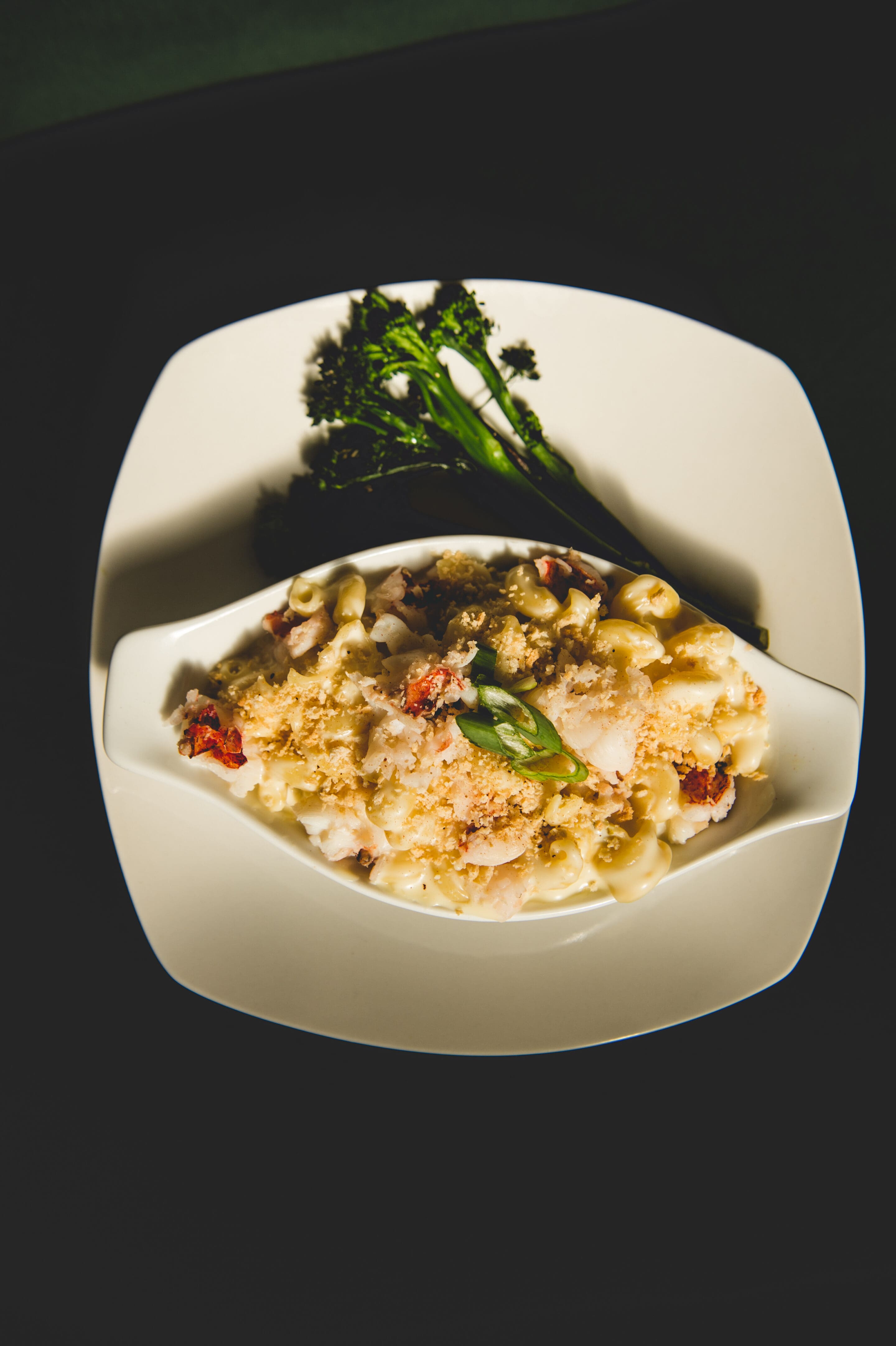 A white dish of baked macaroni and cheese with breadcrumbs and herbs, garnished with green onions, served on a white plate with a side of broccolini.