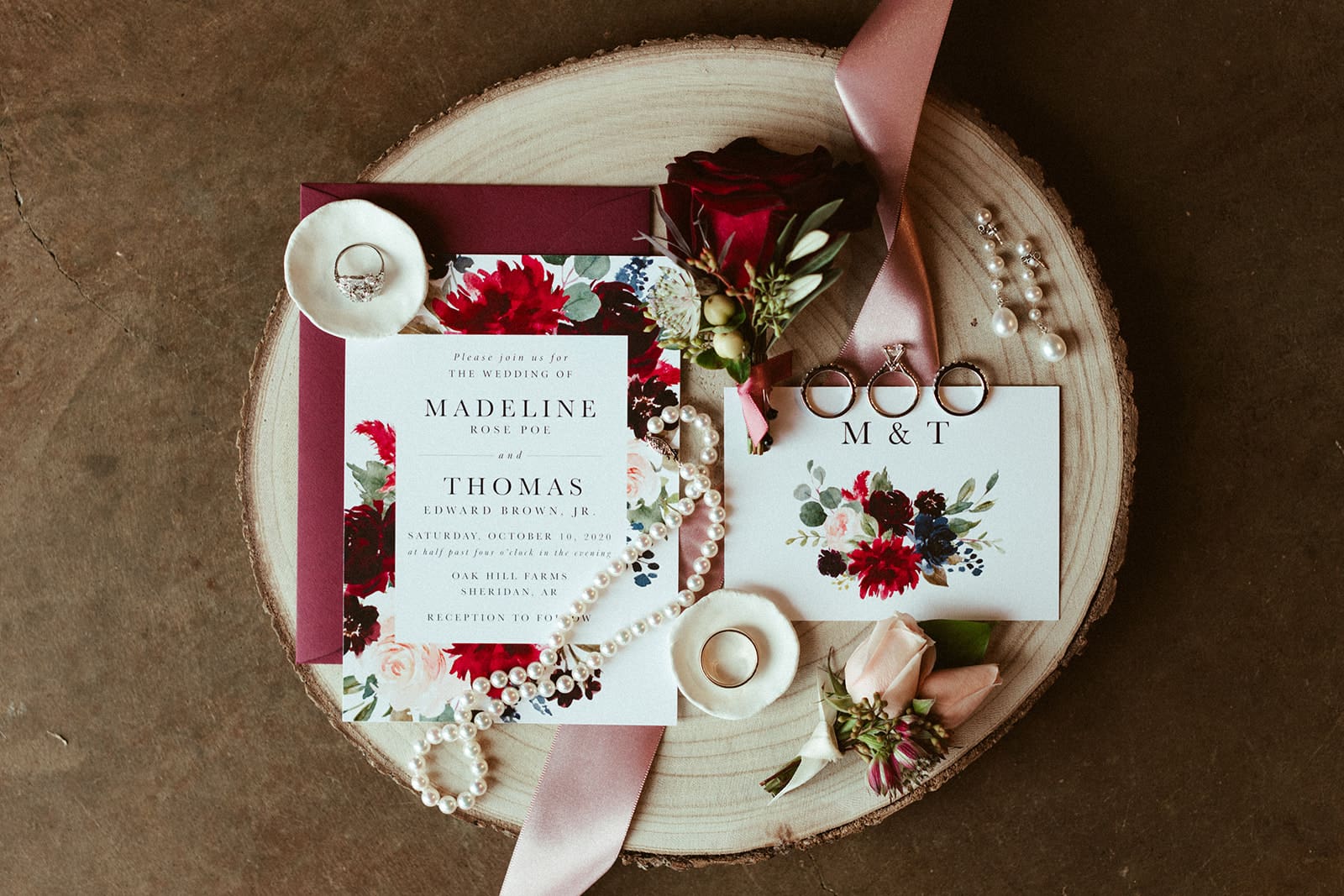 A flat lay of wedding invitations and RSVP card with red and pink floral designs, surrounded by rings, a pearl necklace, flowers, a pink ribbon, and a small dish on a wooden surface.