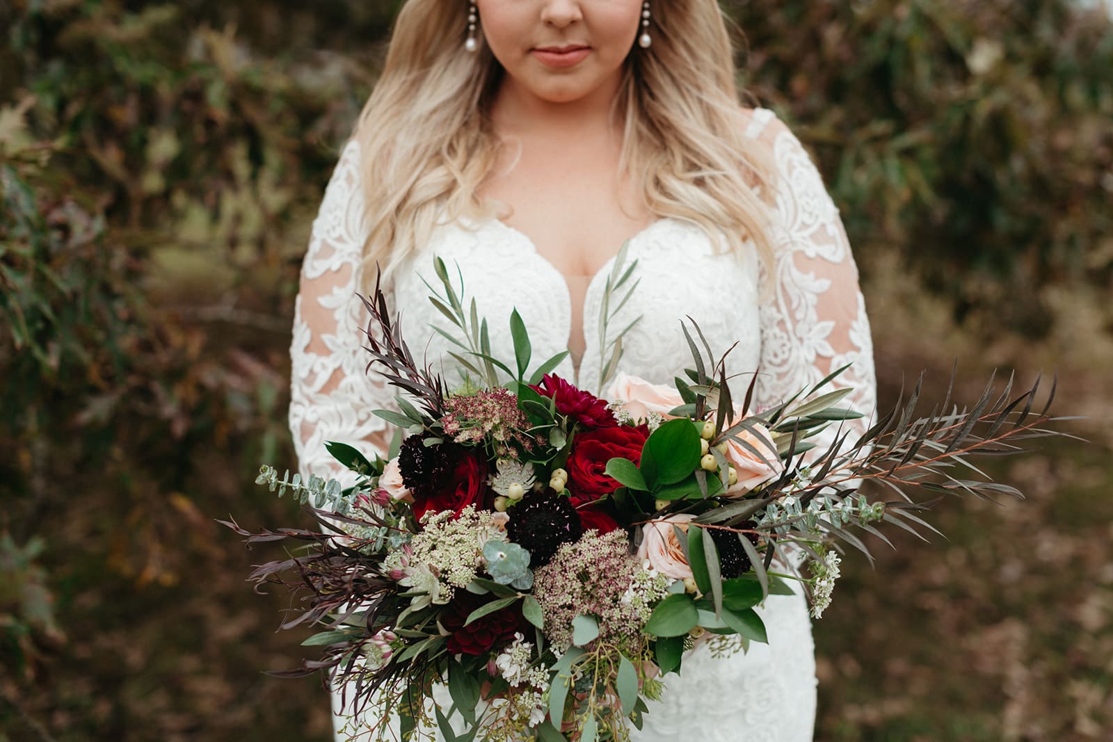 A bride in a lace wedding dress holds a vibrant bouquet with red, pink, and dark flowers, and green foliage, outdoors with greenery in the background. Her face is partially visible.