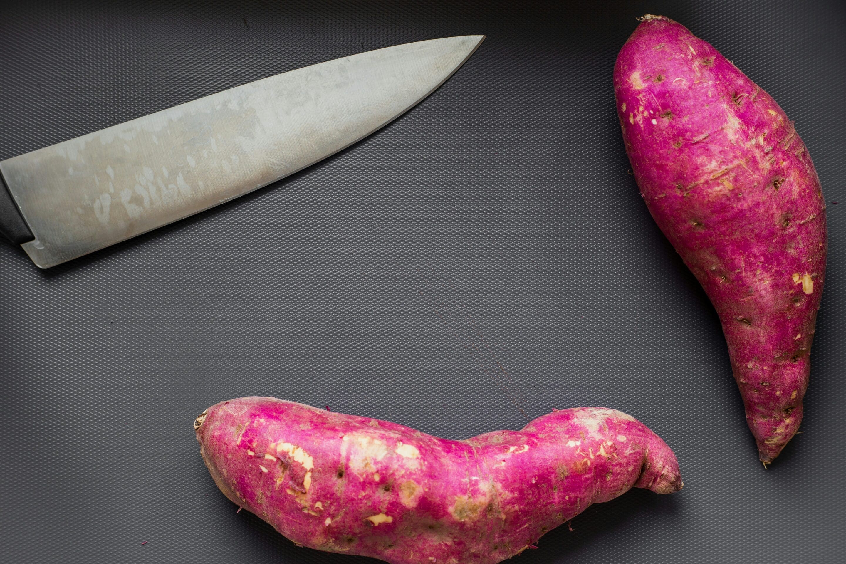 A chef’s knife and two raw purple sweet potatoes are placed on a dark textured surface.