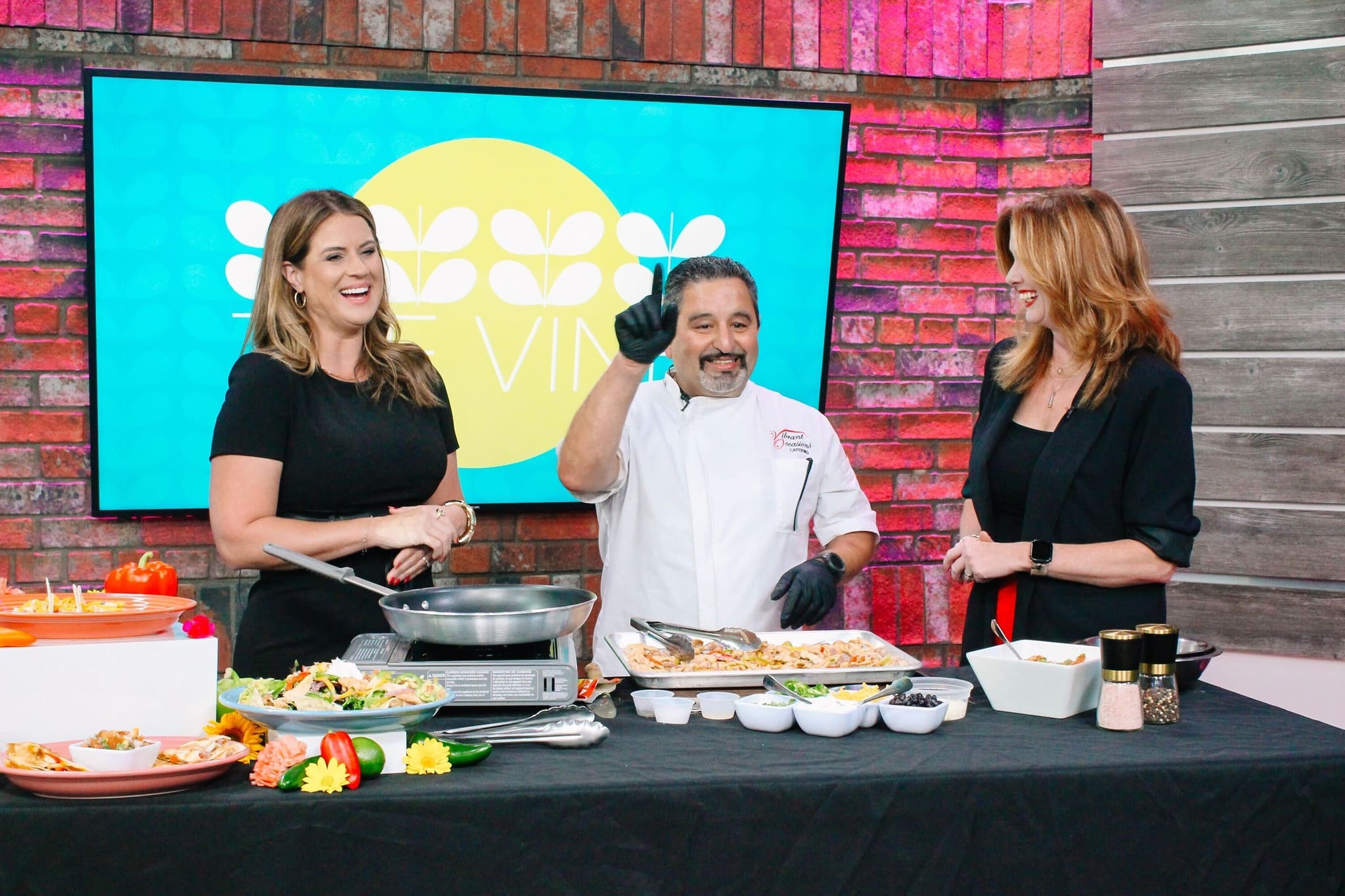 Three people stand behind a table with various dishes, smiling and talking. The man in the center wears a white chefs coat and black gloves. Two women stand on either side. A colorful set with a Viva sign is in the background.