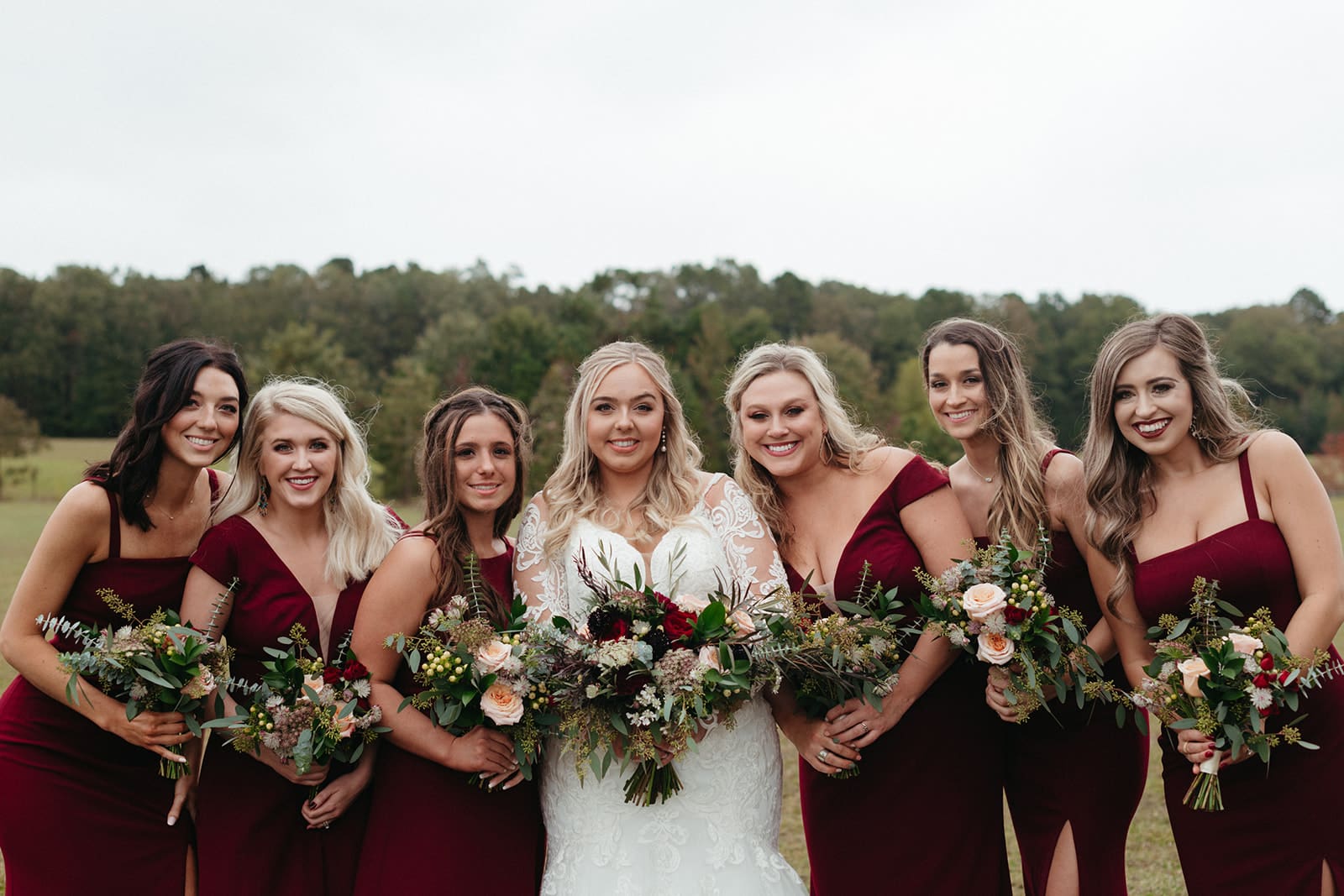 A bride in a white dress stands outdoors with six bridesmaids in matching burgundy dresses, all holding bouquets of flowers and smiling at the camera. Trees and greenery are visible in the background.