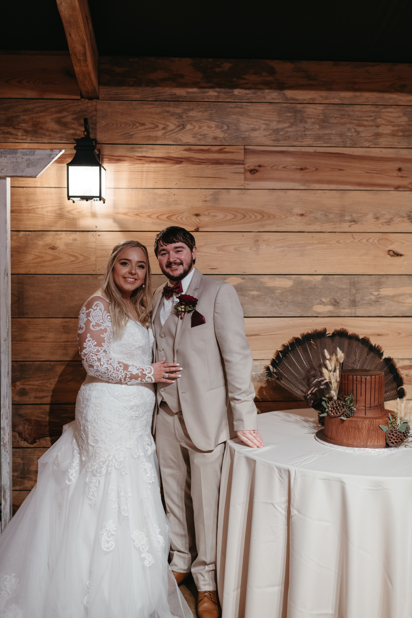 A bride in a white lace gown and a groom in a beige suit stand smiling beside a round table with a rustic brown wedding cake, against a wooden-paneled wall.