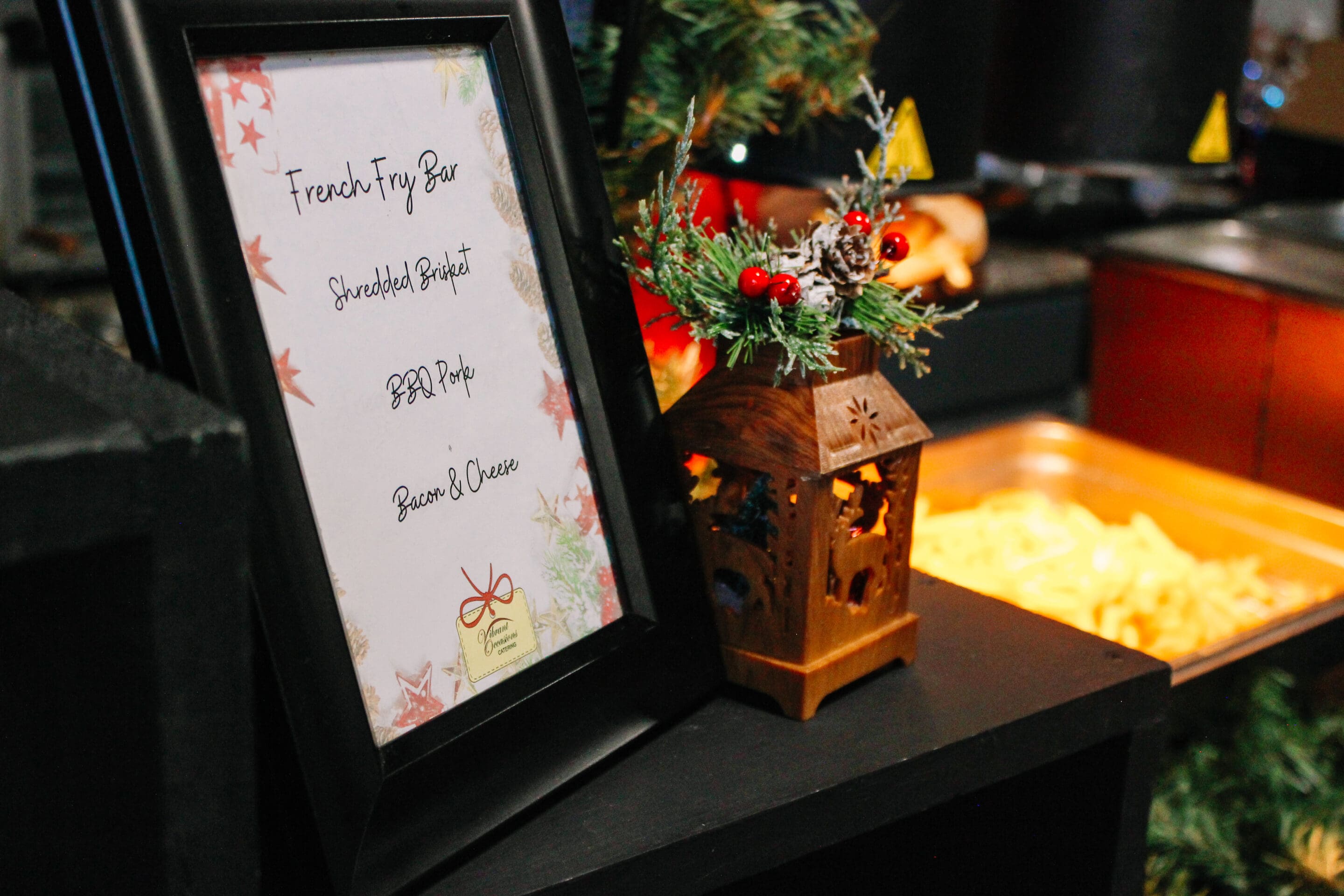 A framed menu reads “French Fry Bar: Shredded Brisket, BBQ Pork, Bacon & Cheese.” Next to it is a small holiday decoration with greenery and berries, and a tray of fries in the background.