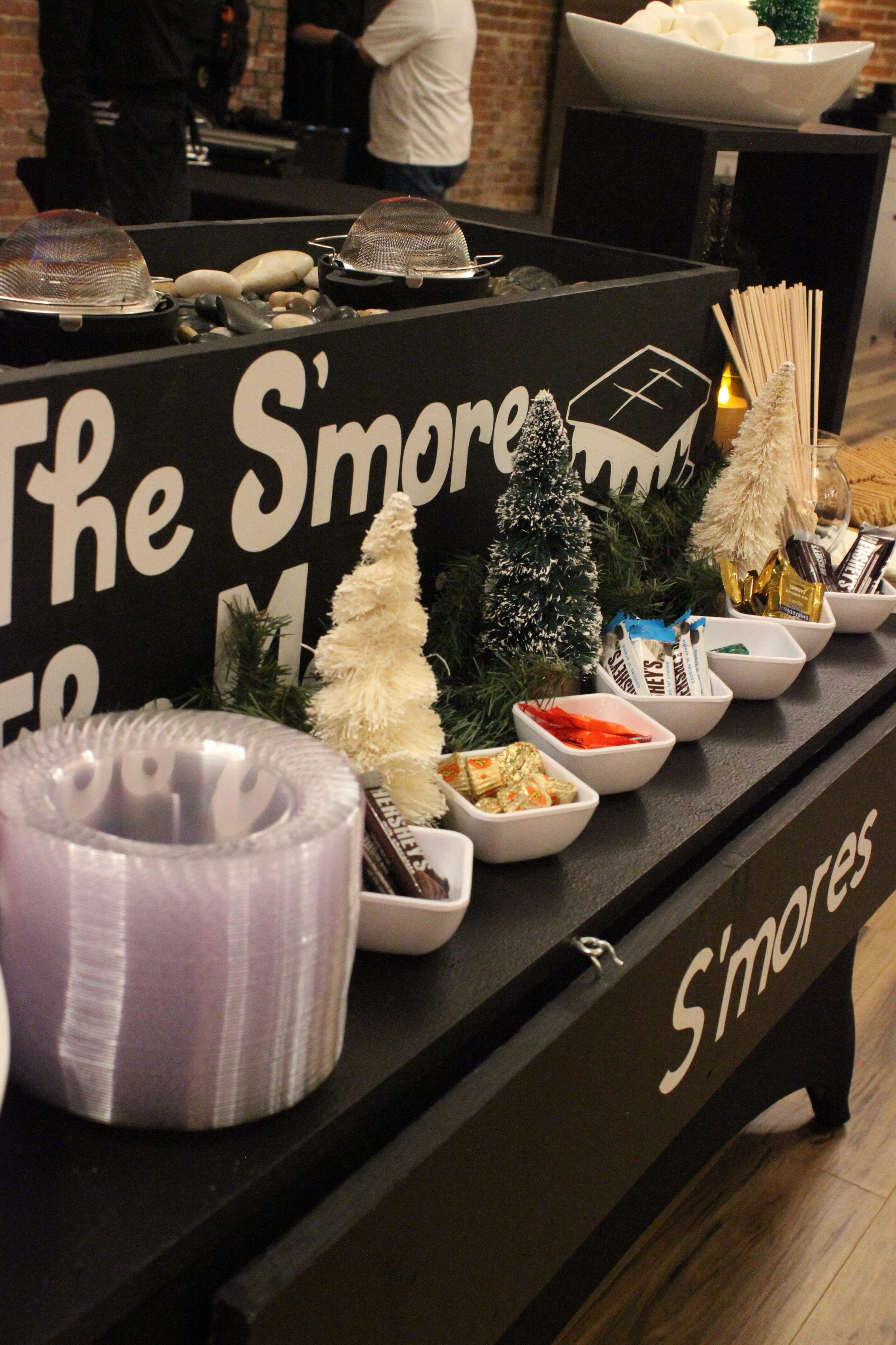 A s’mores station featuring small white bowls filled with candy, chocolate, and graham crackers, decorative mini trees, plates, skewers, and a black sign that reads “The S’mores” and “S’mores.”.