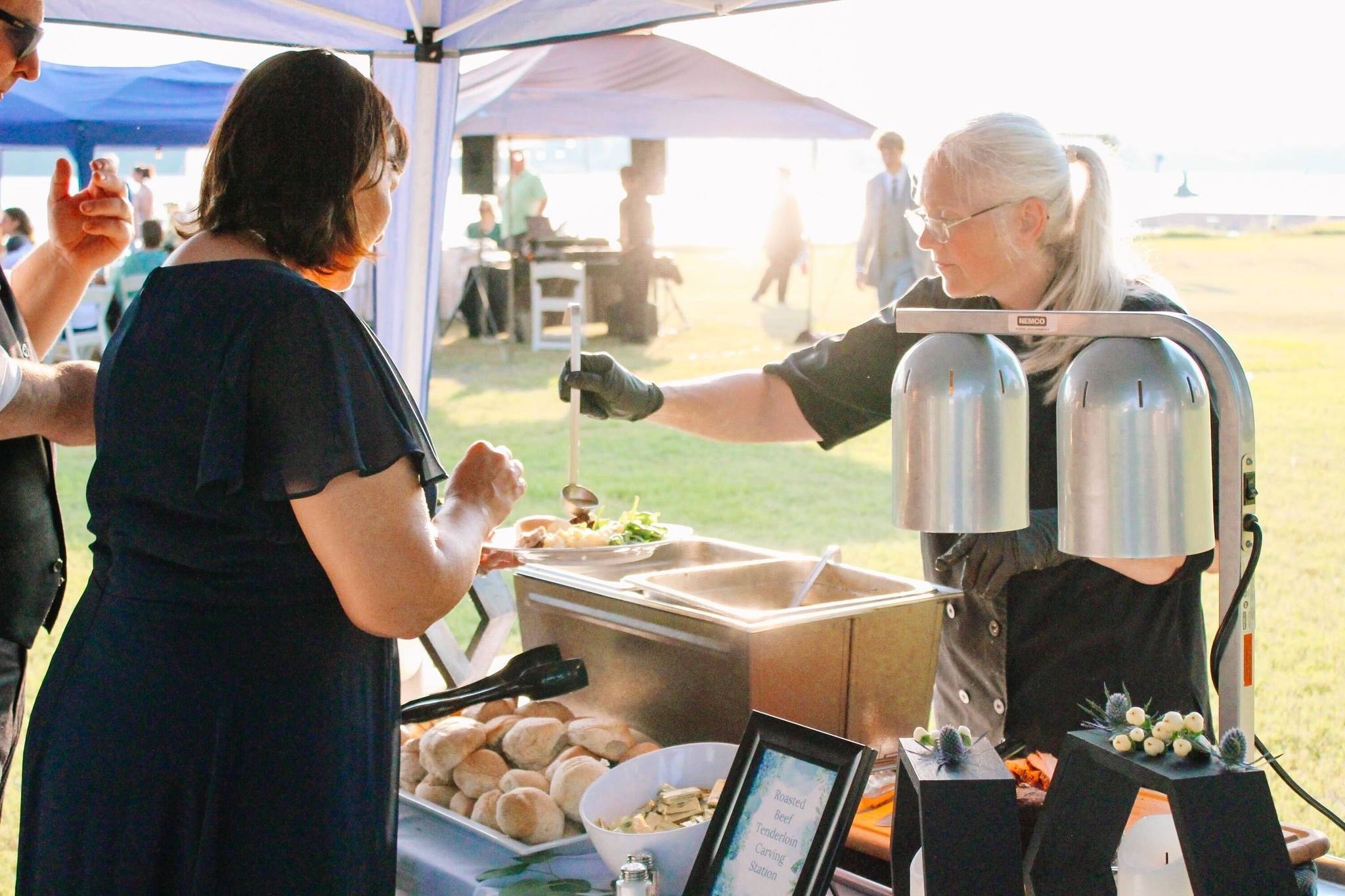 A woman serves food from a buffet station under a canopy at an outdoor event, handing a spoonful to a guest holding a plate. Bread rolls and salad are visible on the table. The event takes place in a sunny, grassy area.