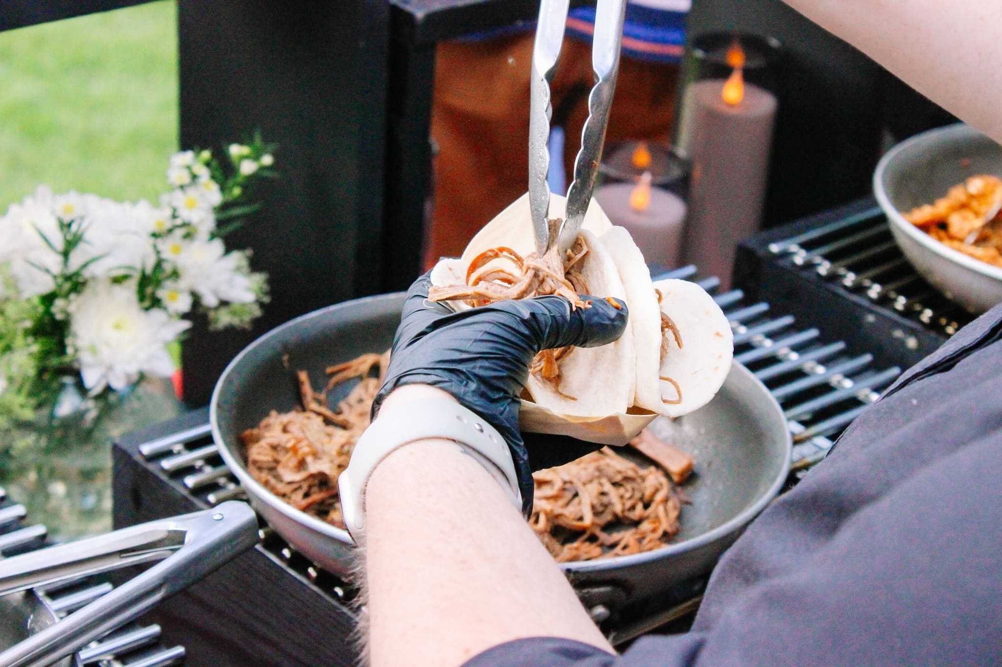 A person wearing black gloves uses tongs to fill a soft taco shell with shredded meat over a pan, with flowers and candles visible in the background.