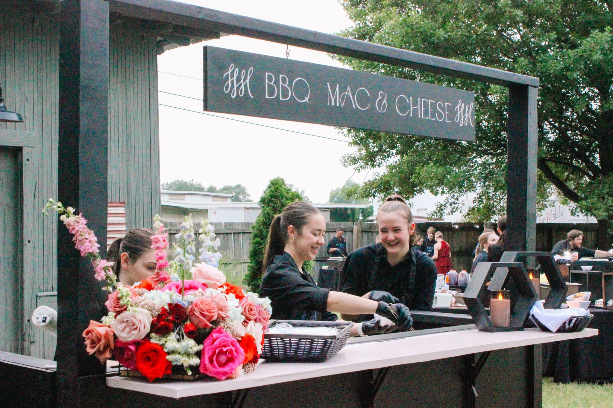 Three women in black uniforms serve food from an outdoor BBQ mac and cheese stand. A bouquet of colorful flowers decorates the counter, and trees are visible in the background. The atmosphere appears festive and welcoming.