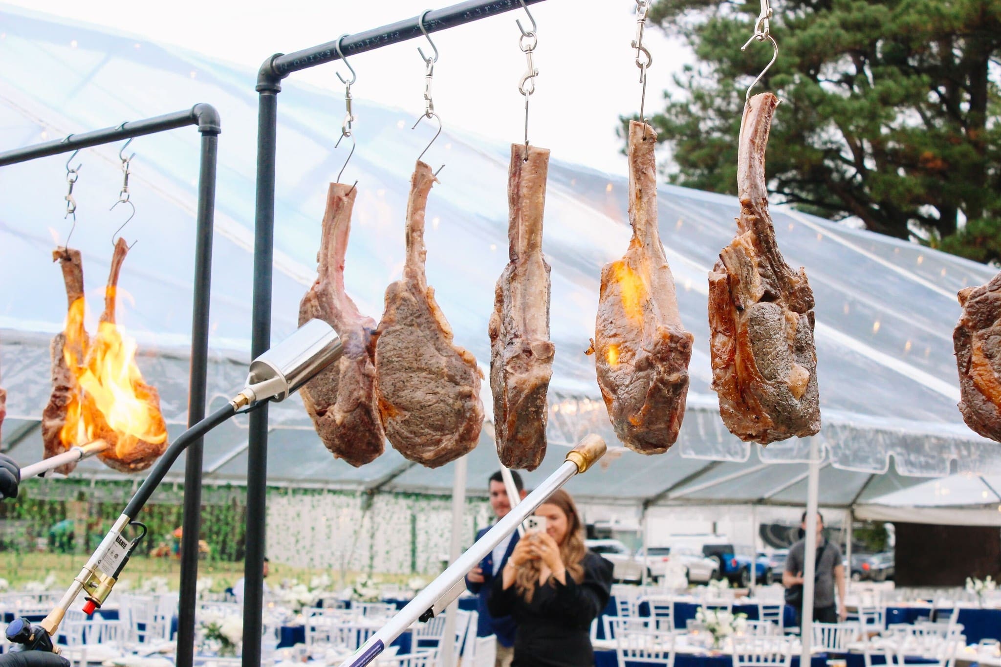 Several large tomahawk steaks hang from metal hooks over an open flame, being seared with a torch. In the background, there are white tables and chairs set up under a clear tent, with people and trees visible.