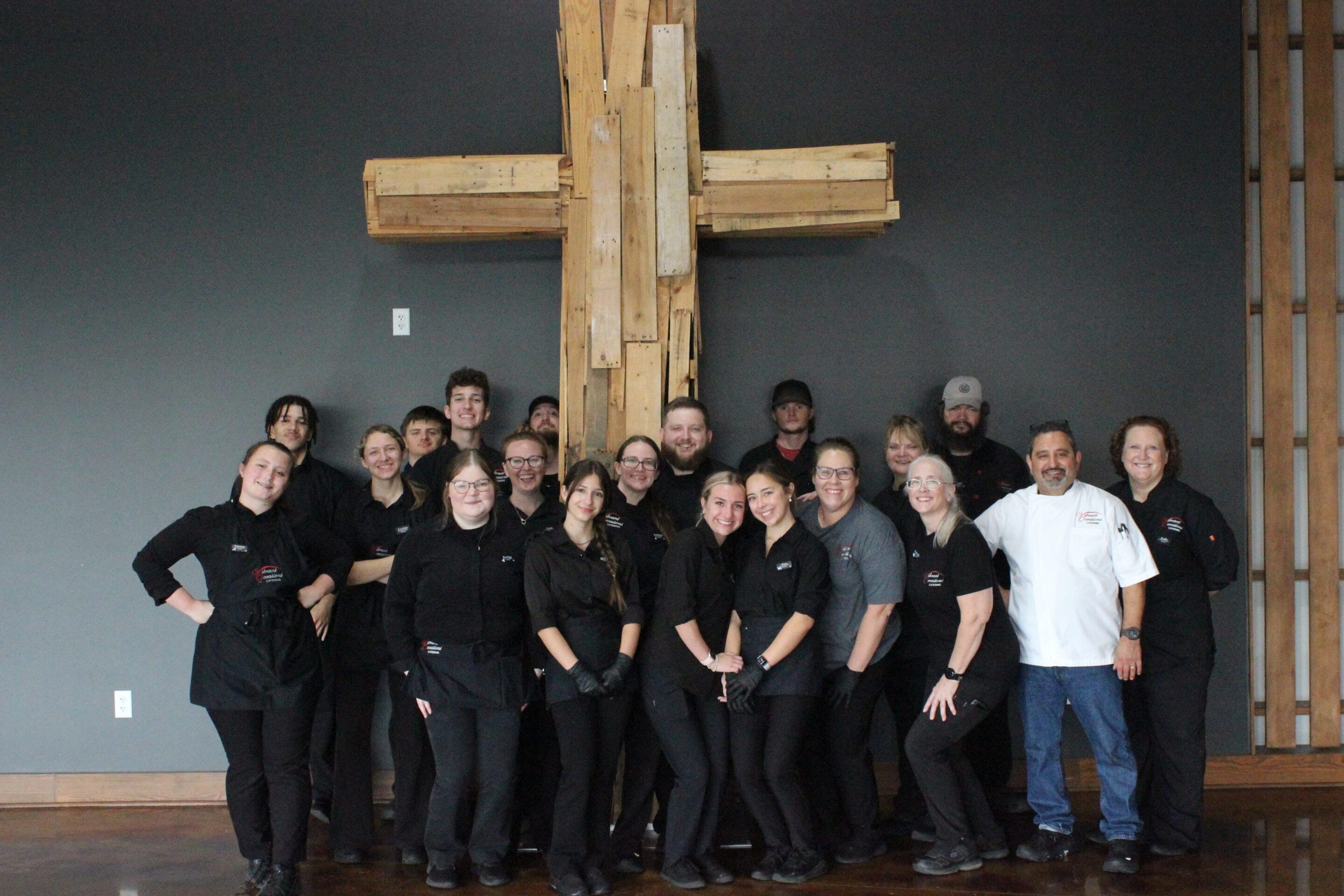 A group of people wearing black uniforms and a few in white chef coats stand smiling in front of a large wooden cross mounted on a gray wall. The group appears to be staff or a team, posing together indoors.