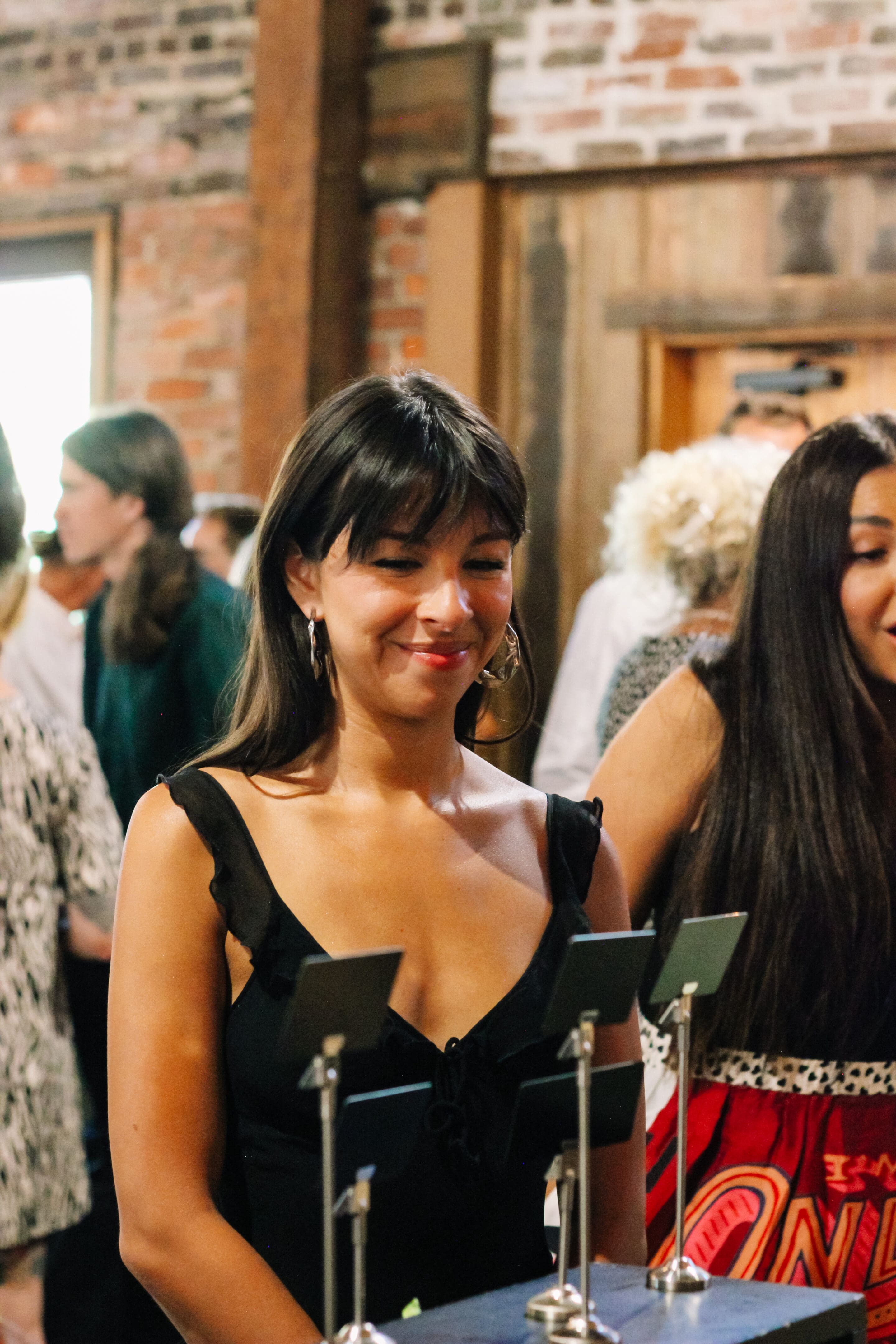 A woman with dark hair and a black dress smiles while looking at display cards on stands, with people and a rustic brick and wood interior in the background.
