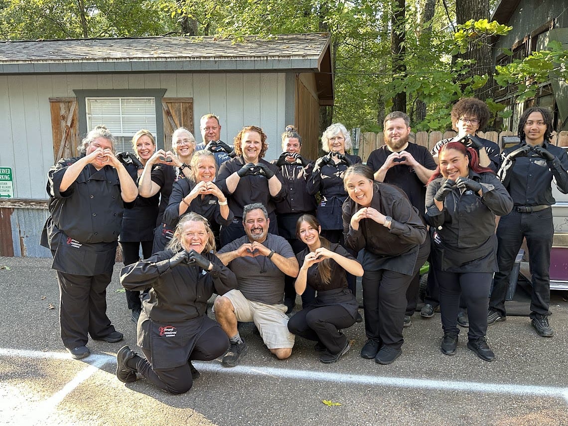 A group of people, mostly dressed in black uniforms, stand and kneel outdoors in front of a shed, smiling and making heart shapes with their hands for the camera. Trees and greenery are visible in the background.