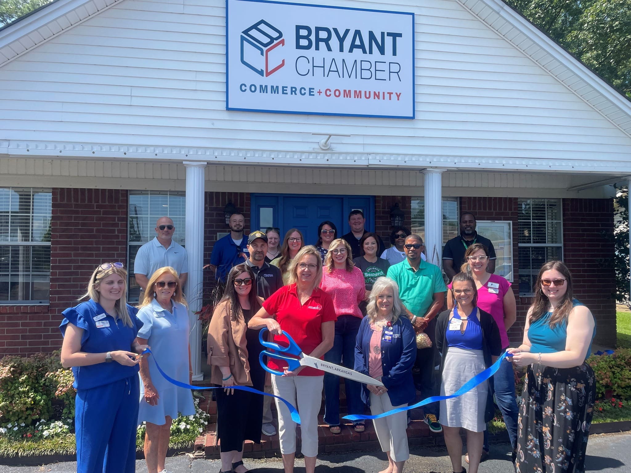 A group of people stand smiling outside the Bryant Chamber building, as a woman cuts a blue ribbon with oversized scissors during an outdoor ribbon-cutting ceremony.
