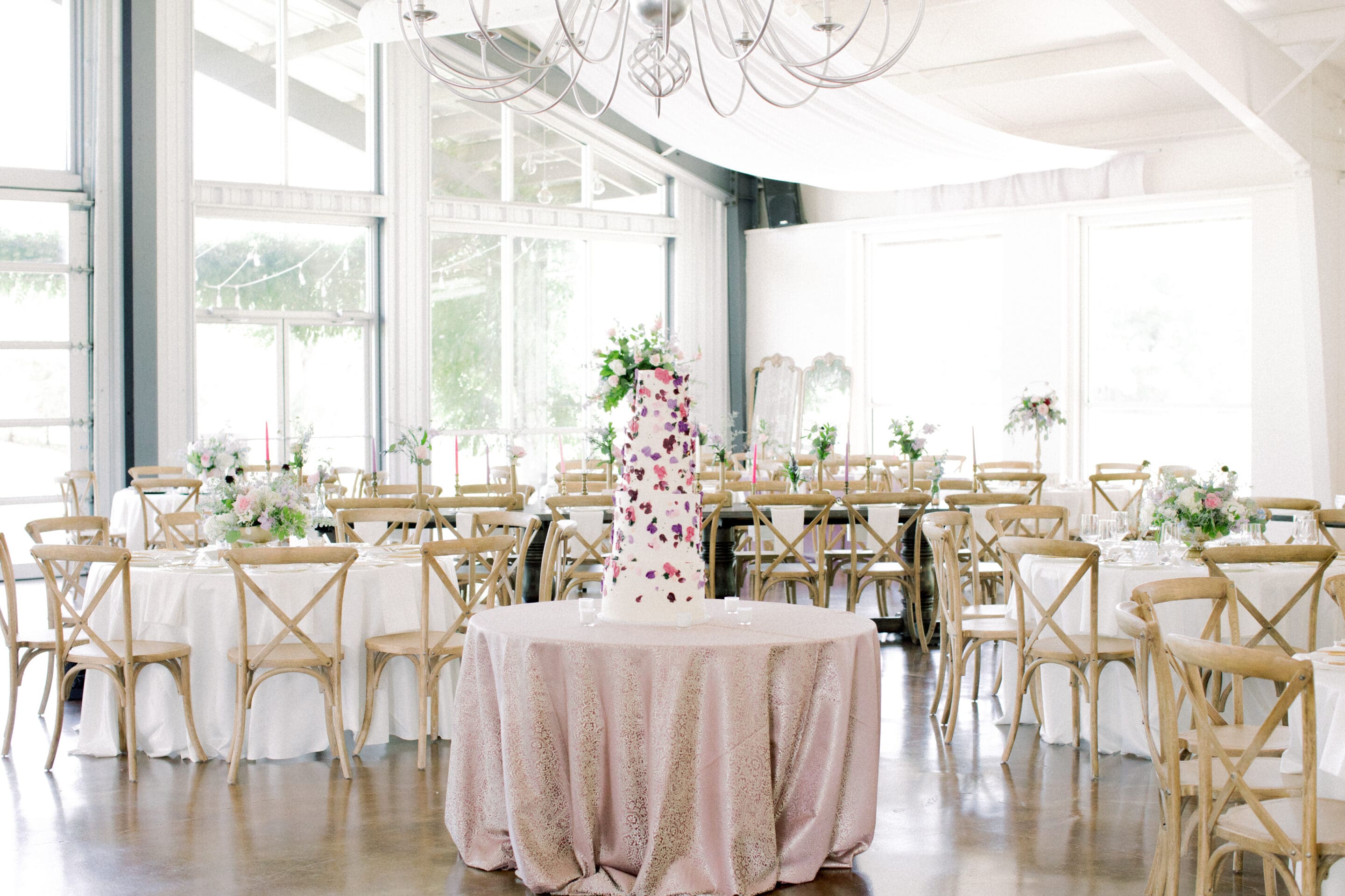 A bright, elegant event space features round tables with white cloths and wooden chairs, floral centerpieces, and a tall, floral-decorated cake on a pink-covered table in the center. Natural light fills the room.