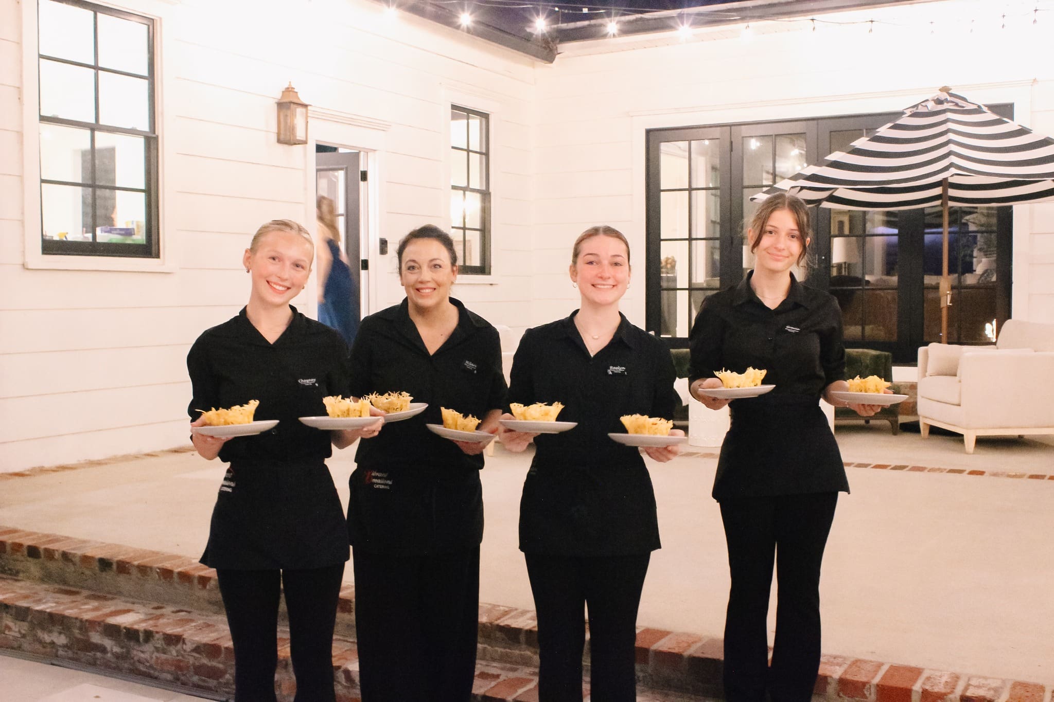 Four smiling servers in black uniforms stand in a row outdoors, each holding plates of food with fries. String lights and patio furniture are visible in the background.