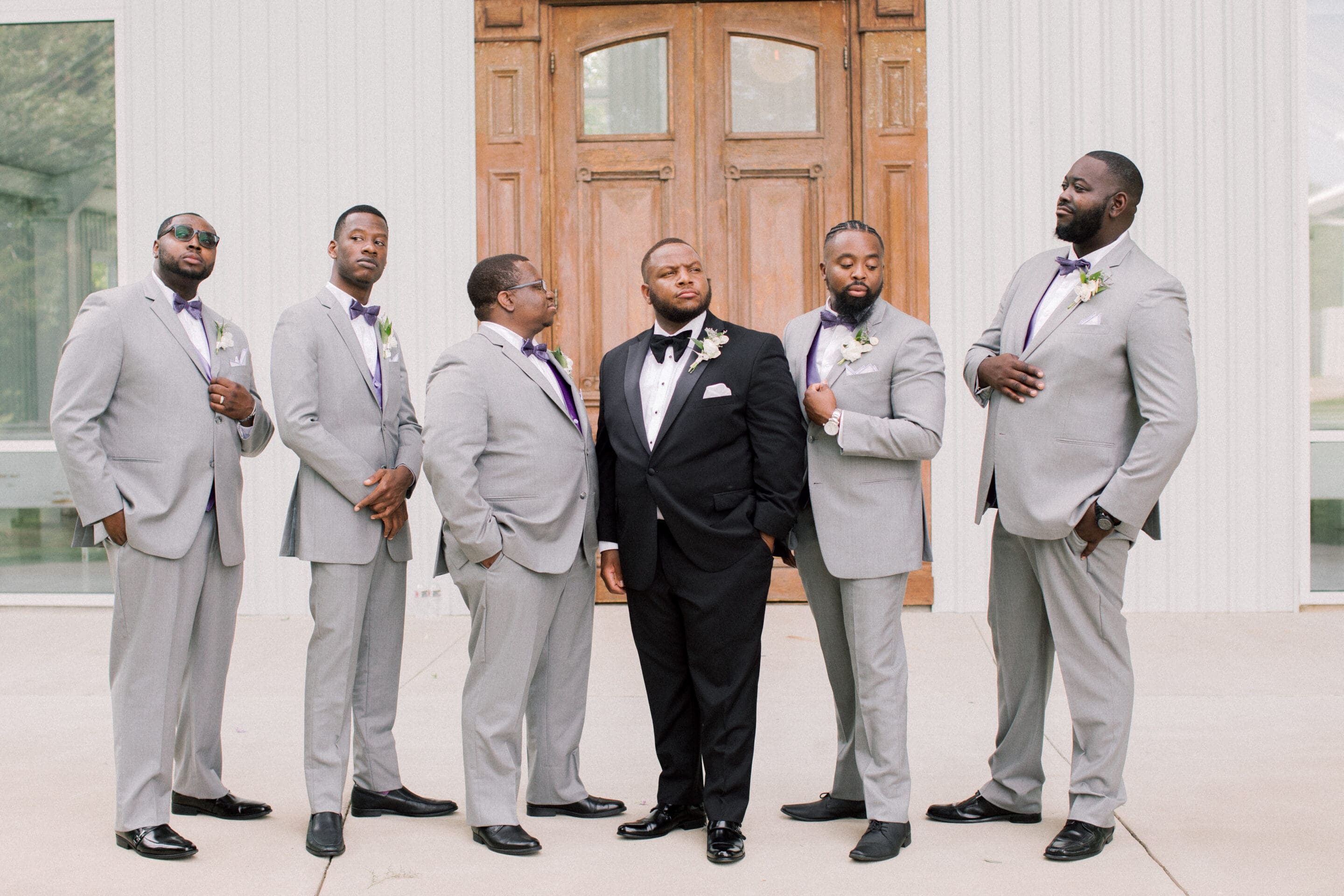 Six men pose in front of a wooden door; five are in light gray suits with purple bow ties, and one, centered, wears a black tuxedo with a bow tie. All look stylish and confident on a formal occasion.