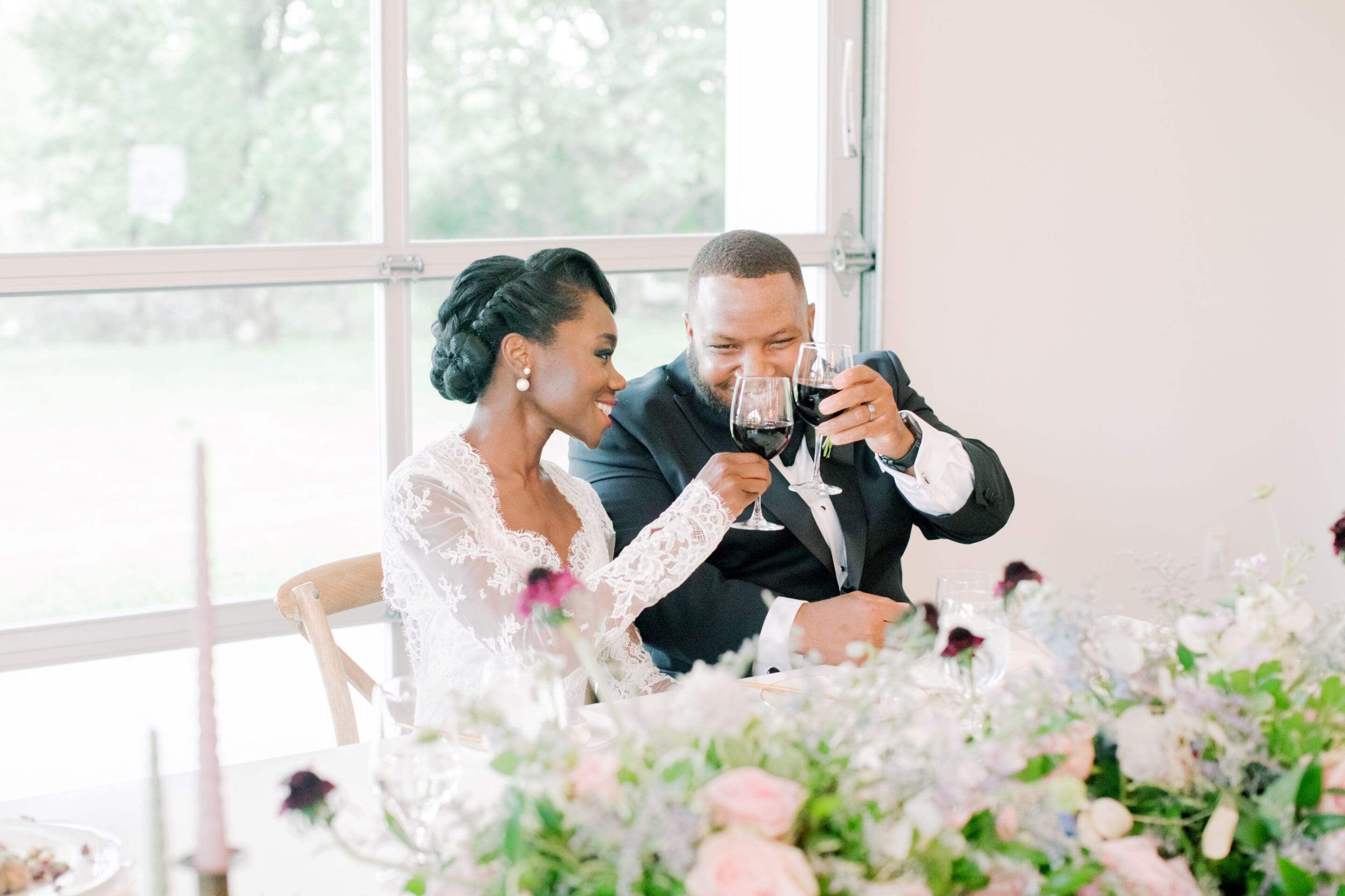 A bride and groom in formal attire sit at a decorated table, smiling and toasting with wine glasses, surrounded by pastel flowers and natural light.