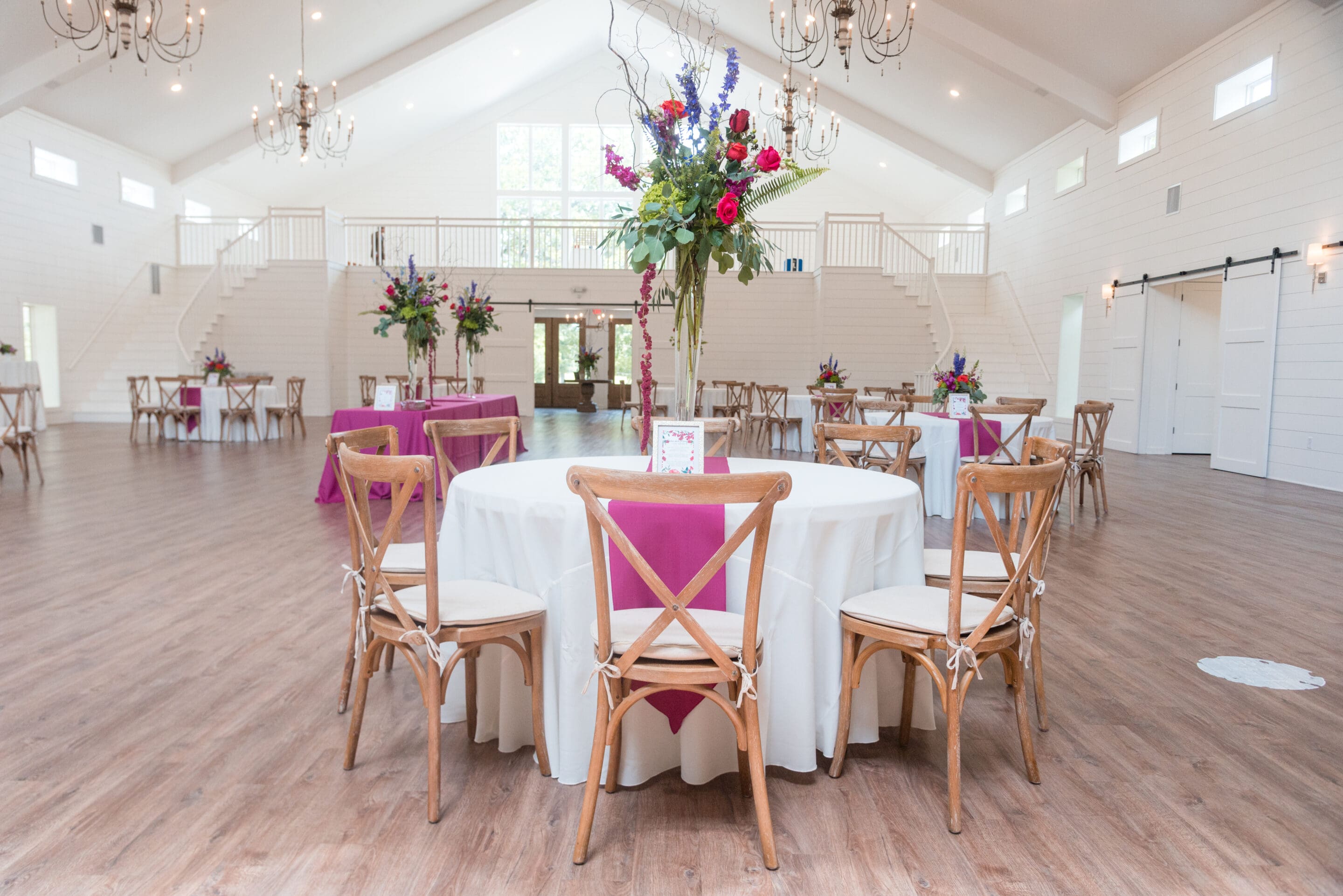 A bright, spacious event hall with white walls and high ceilings features round tables draped in white tablecloths, wooden chairs, and tall floral centerpieces with pink, purple, and green arrangements.