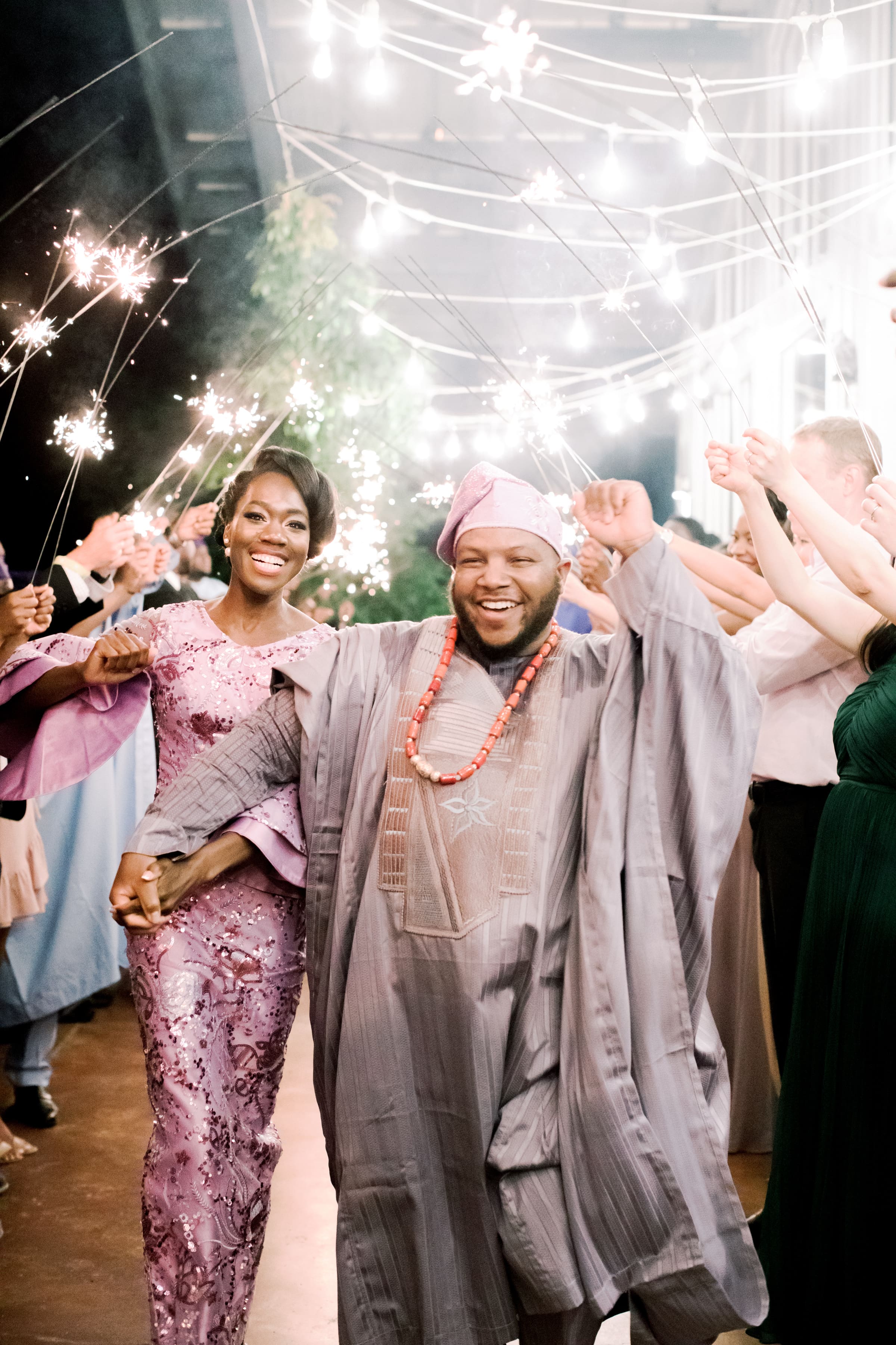 A joyful couple in traditional Nigerian attire walks hand in hand through a crowd holding sparklers, celebrating their wedding. The groom wears agbada and a cap, while the bride wears a pink dress and smiles brightly.