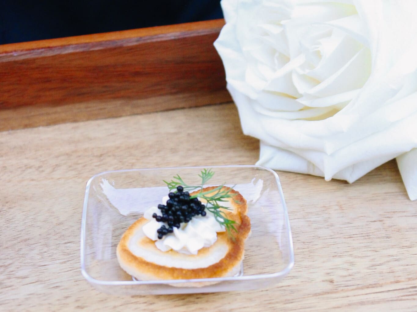 A small appetizer with whipped cream, black caviar, and dill sits in a clear dish on a wooden surface next to a large white rose.