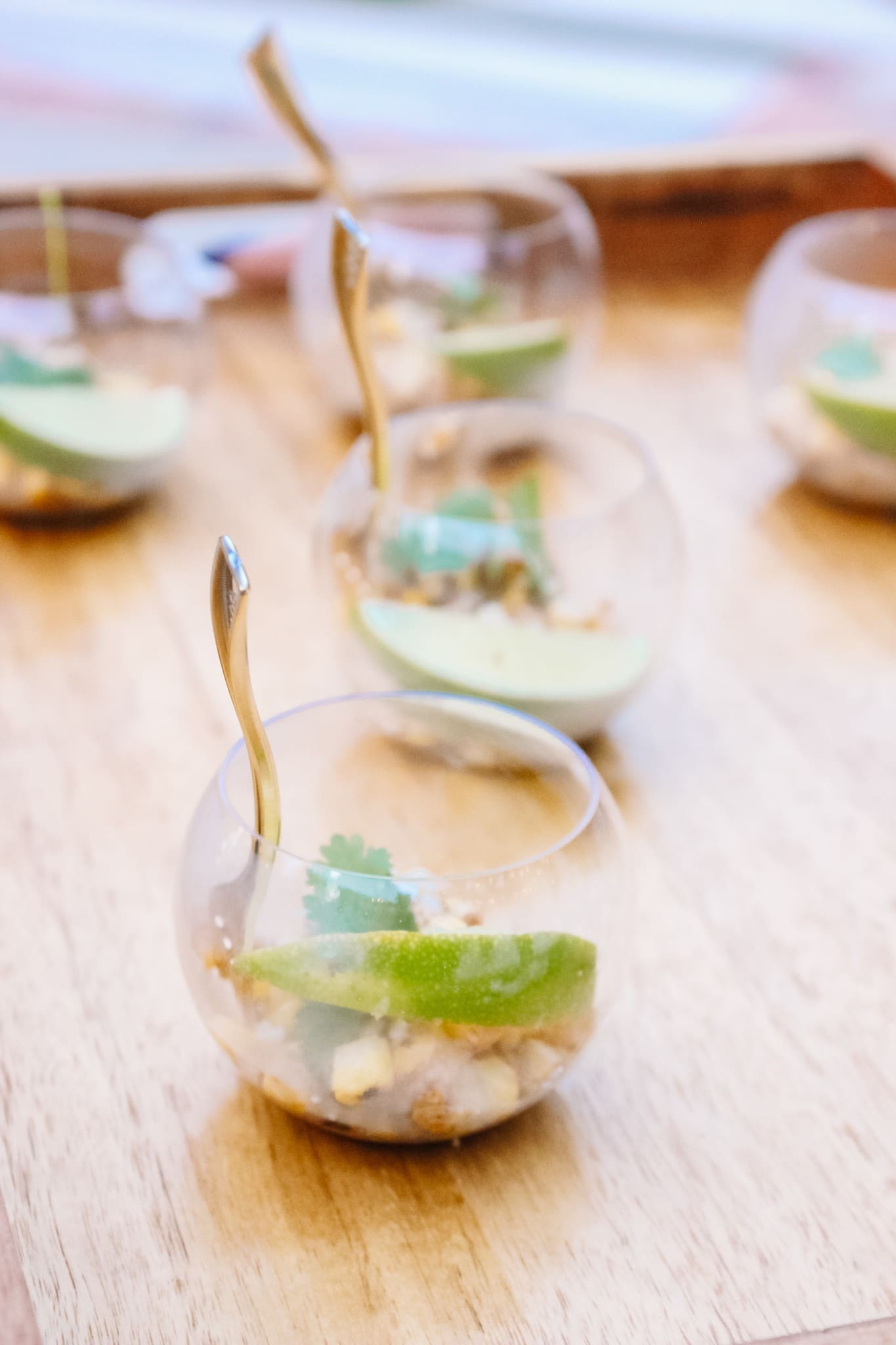 Five small glass cups on a wooden tray, each containing a dessert or appetizer garnished with a lime wedge, cilantro, and nuts, with a gold spoon resting inside each cup.