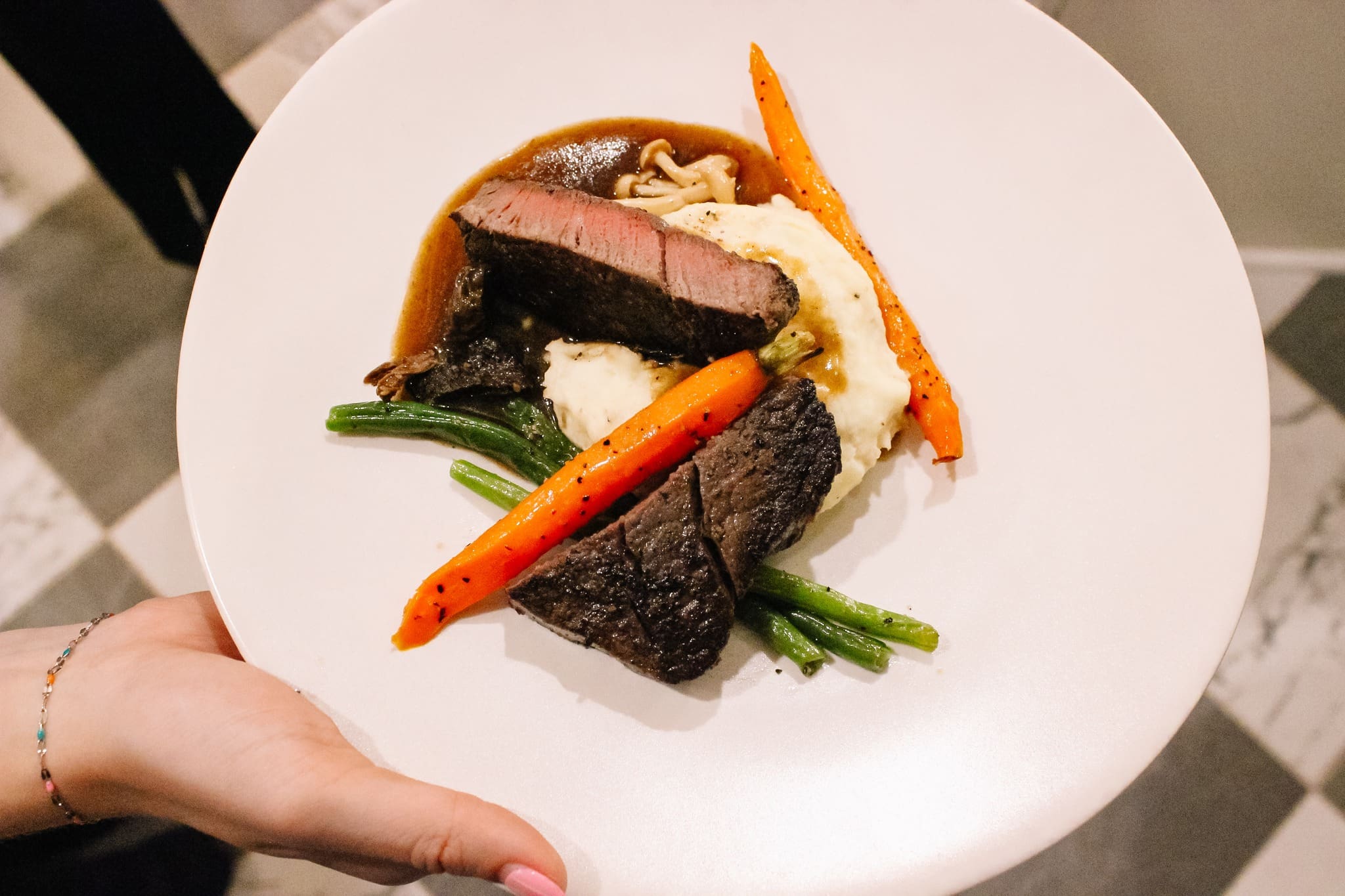 A white plate with slices of medium-rare steak, mashed potatoes, green beans, glazed carrots, and brown sauce, held by a person with a beaded bracelet.