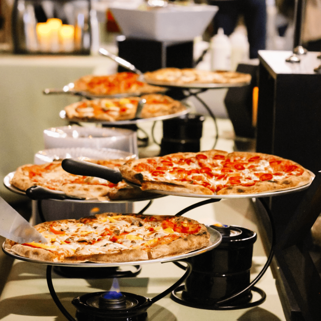 Several pizzas are displayed on metal stands with serving utensils at a buffet table, with each pizza featuring different toppings. In the background, there are blurred candles and serving dishes.