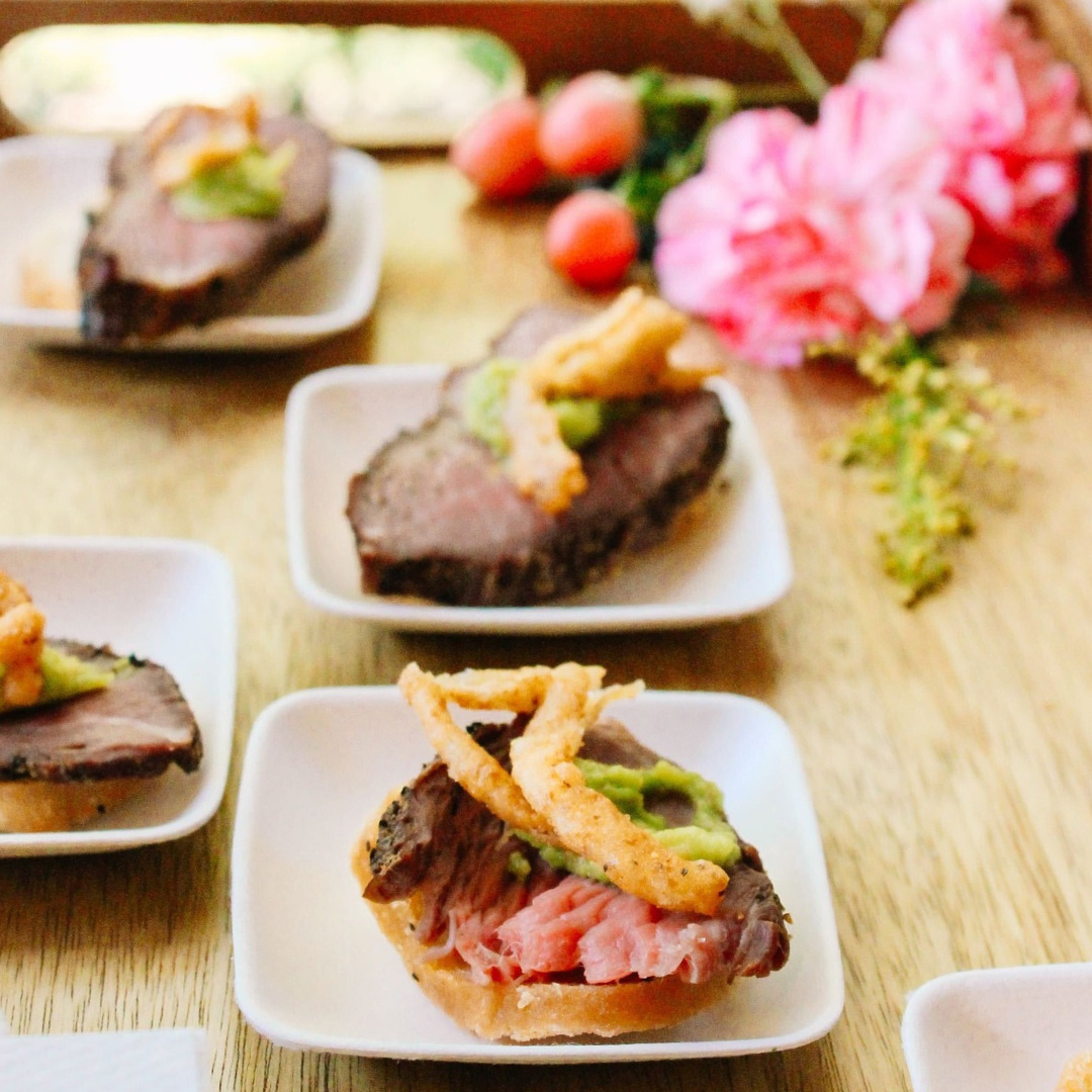 Small plates of sliced roast beef topped with crispy onions and green sauce are arranged on a wooden surface, with pink flowers and small tomatoes in the background.
