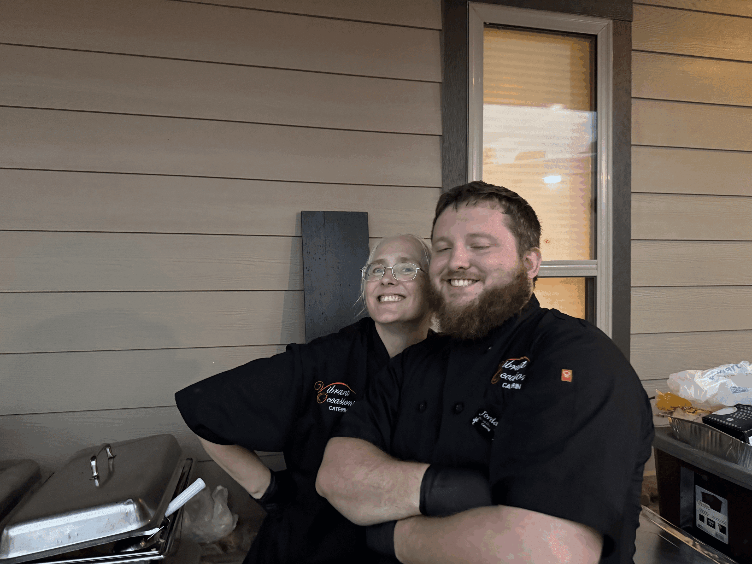 Two smiling chefs in black uniforms stand close together outside in front of a beige wall and window. One chef has glasses and hands on their hips, while the other stands with arms crossed. Catering trays are visible beside them.