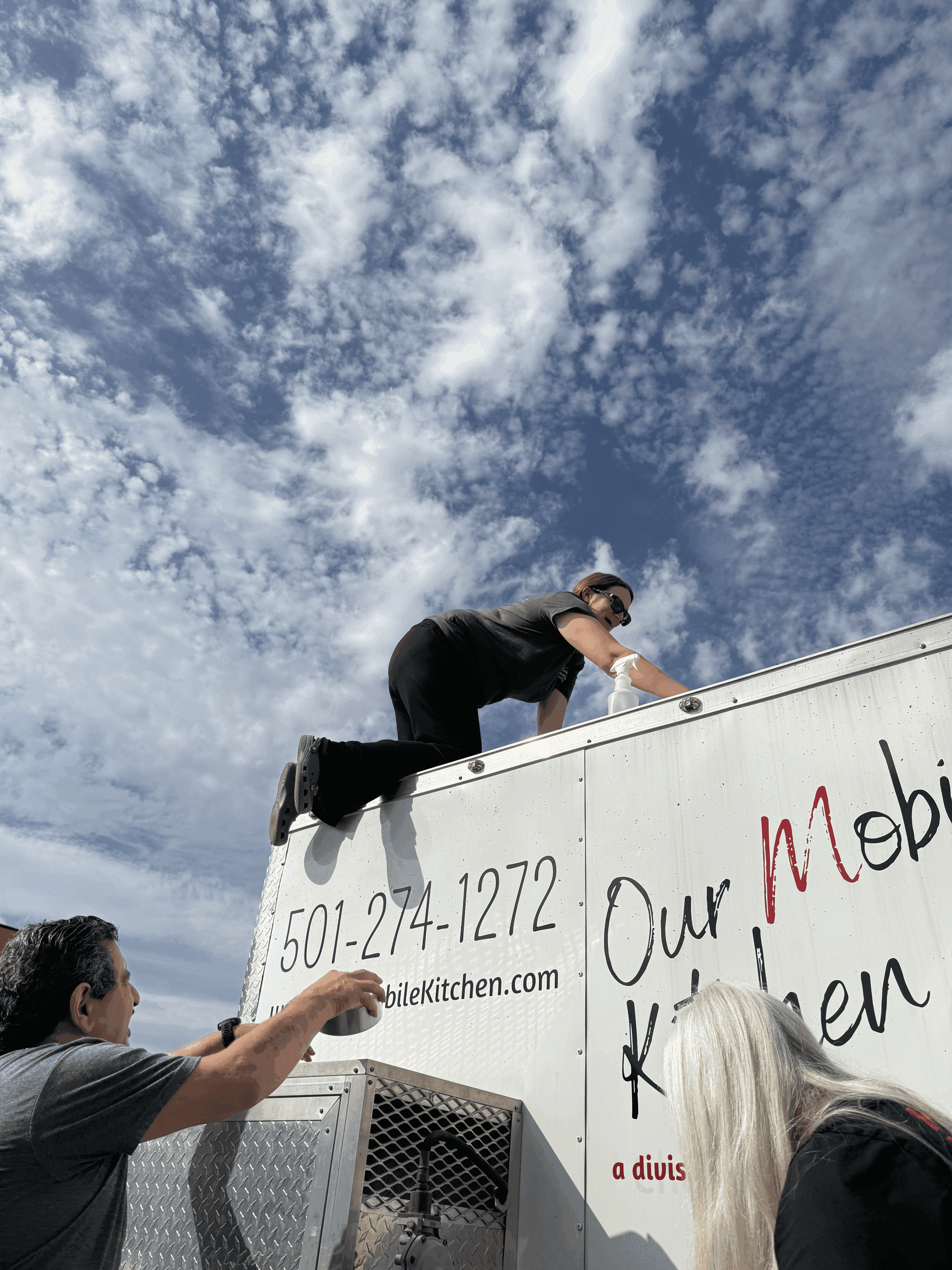 A man in sunglasses climbs on top of a white food truck as two other people watch from below. The sky is partly cloudy with scattered white clouds. The side of the truck displays a phone number and the words Our Mobile Kitchen.