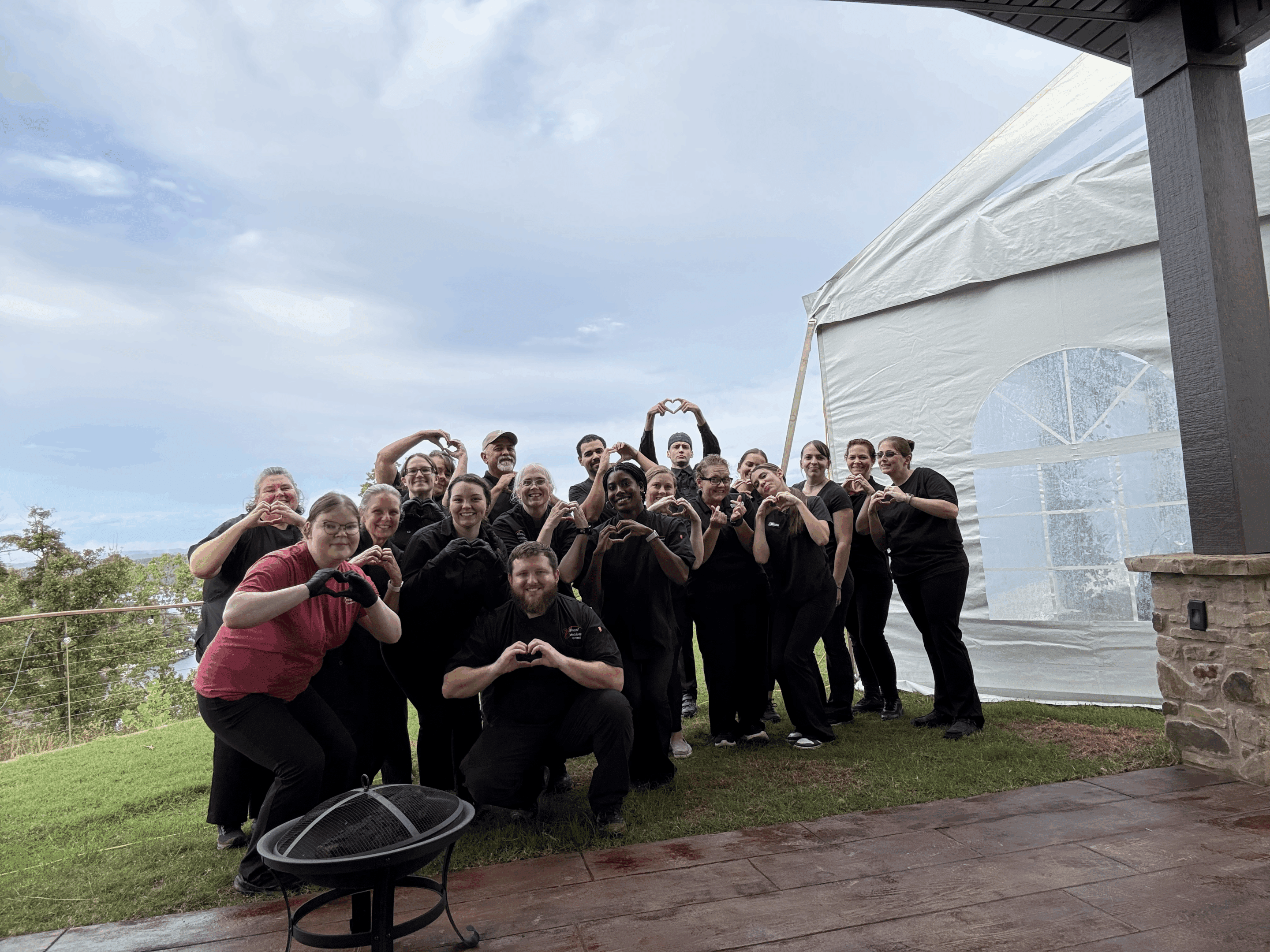 A group of people dressed in black, some with aprons, smile and form heart shapes with their hands while standing on a patio near a large white tent, with trees and a cloudy sky in the background.