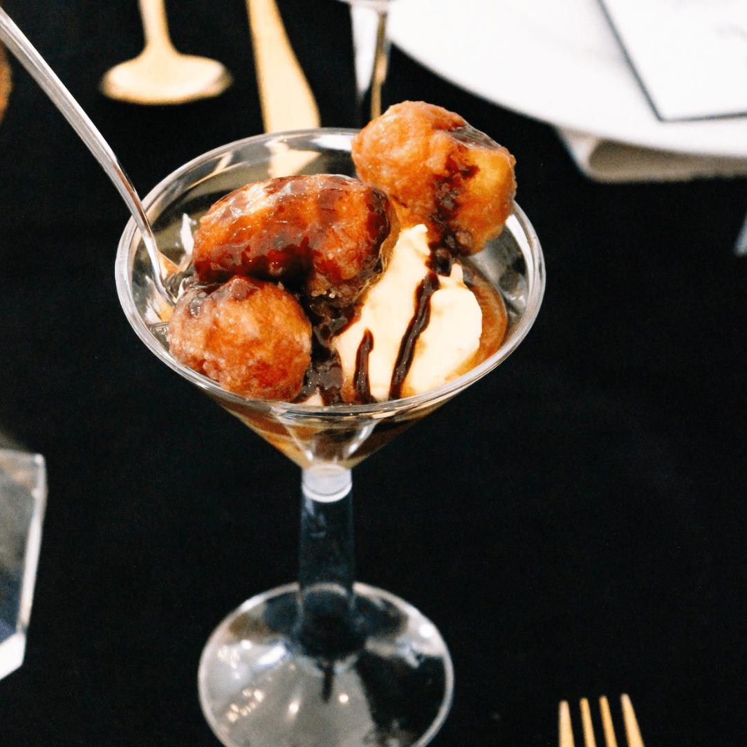 A martini glass filled with vanilla ice cream, fried dough balls, and chocolate sauce, with a spoon placed inside. The background features a black tablecloth and blurred cutlery.