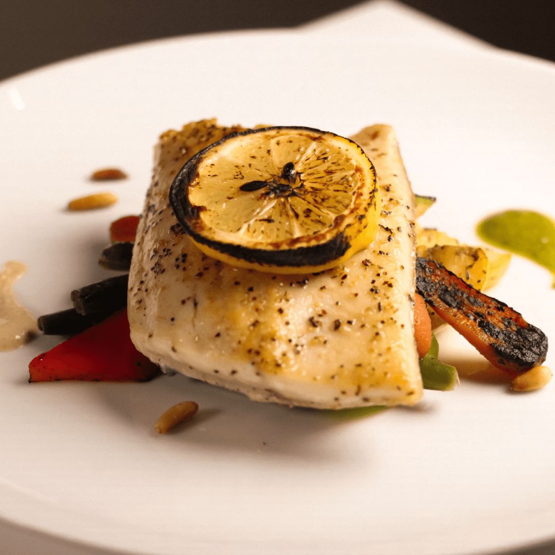 A fillet of fish seasoned with pepper, topped with a grilled lemon slice, served on roasted vegetables and garnished with sauce on a white plate.